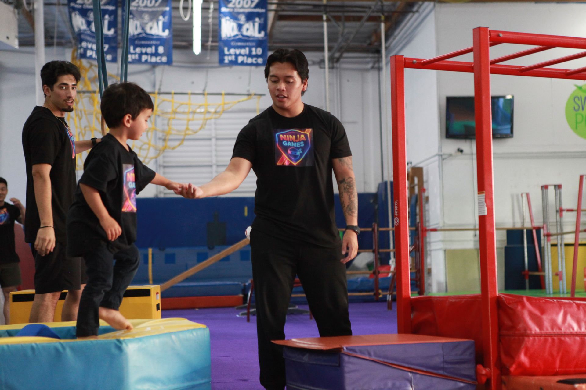 Child and two adults in a gym, child reaching for a hand. Gym equipment visible.