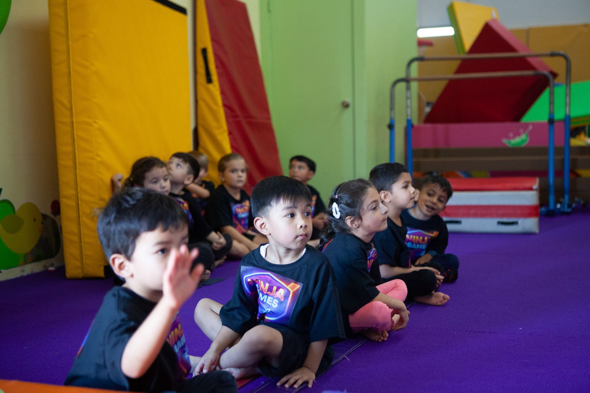 Children sitting in a gymnastics gym, watching; one waves. Purple floor, mats, equipment.