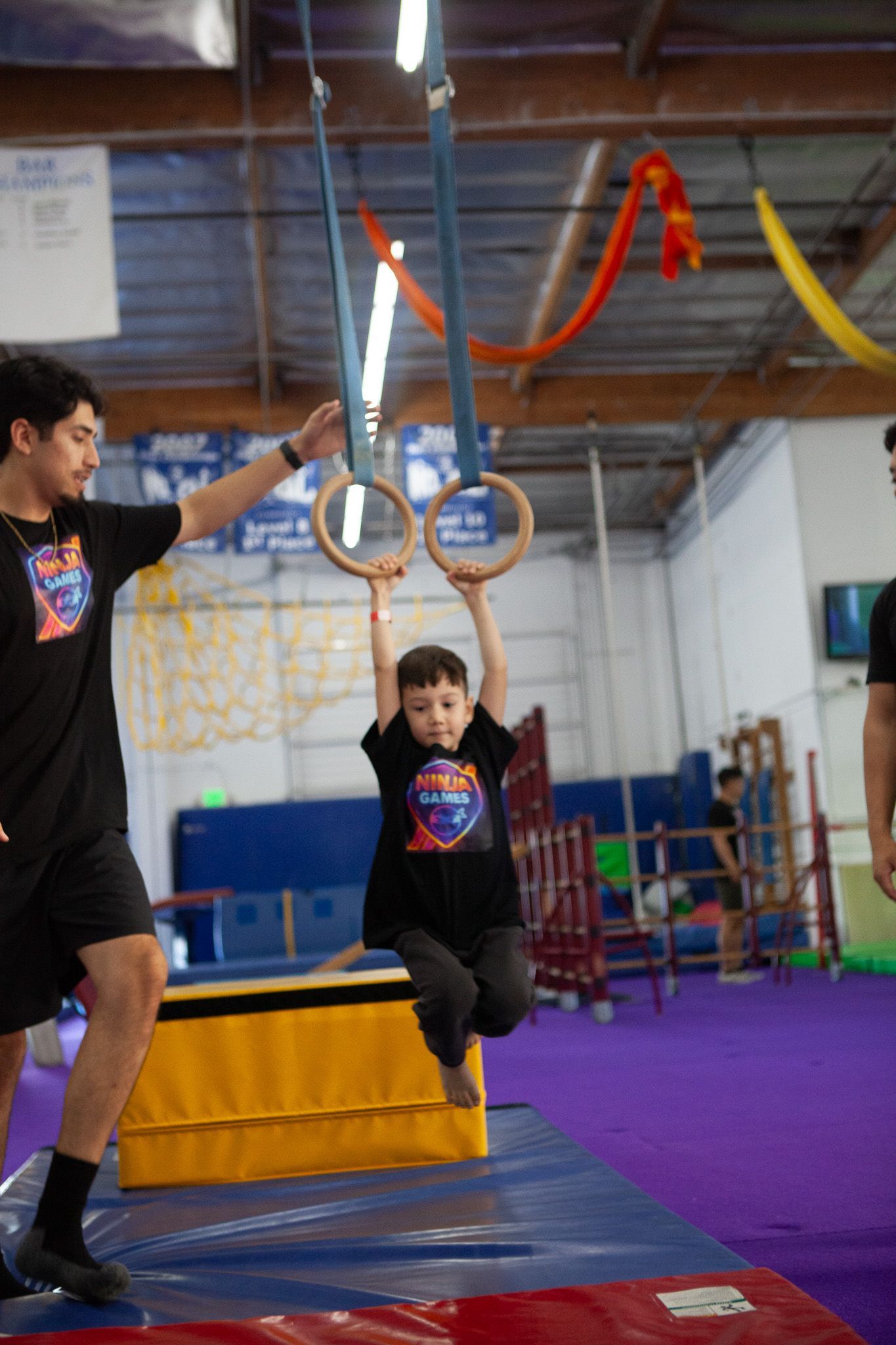Young child hanging from gymnastic rings, instructor spotting. Gym setting with mats, purple floor.