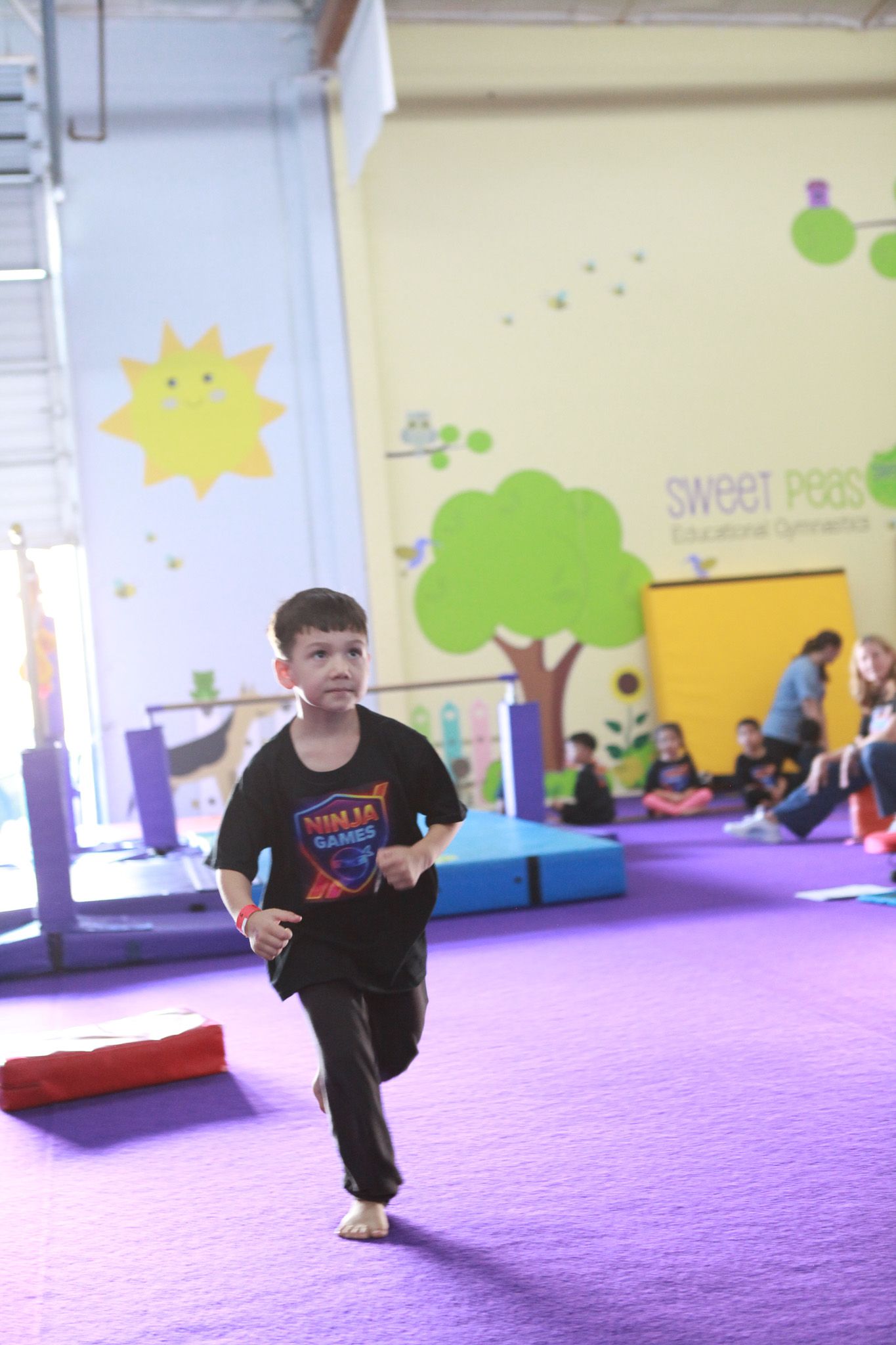 Boy running inside a gymnastics gym with purple mat. Yellow and green wall decorations.