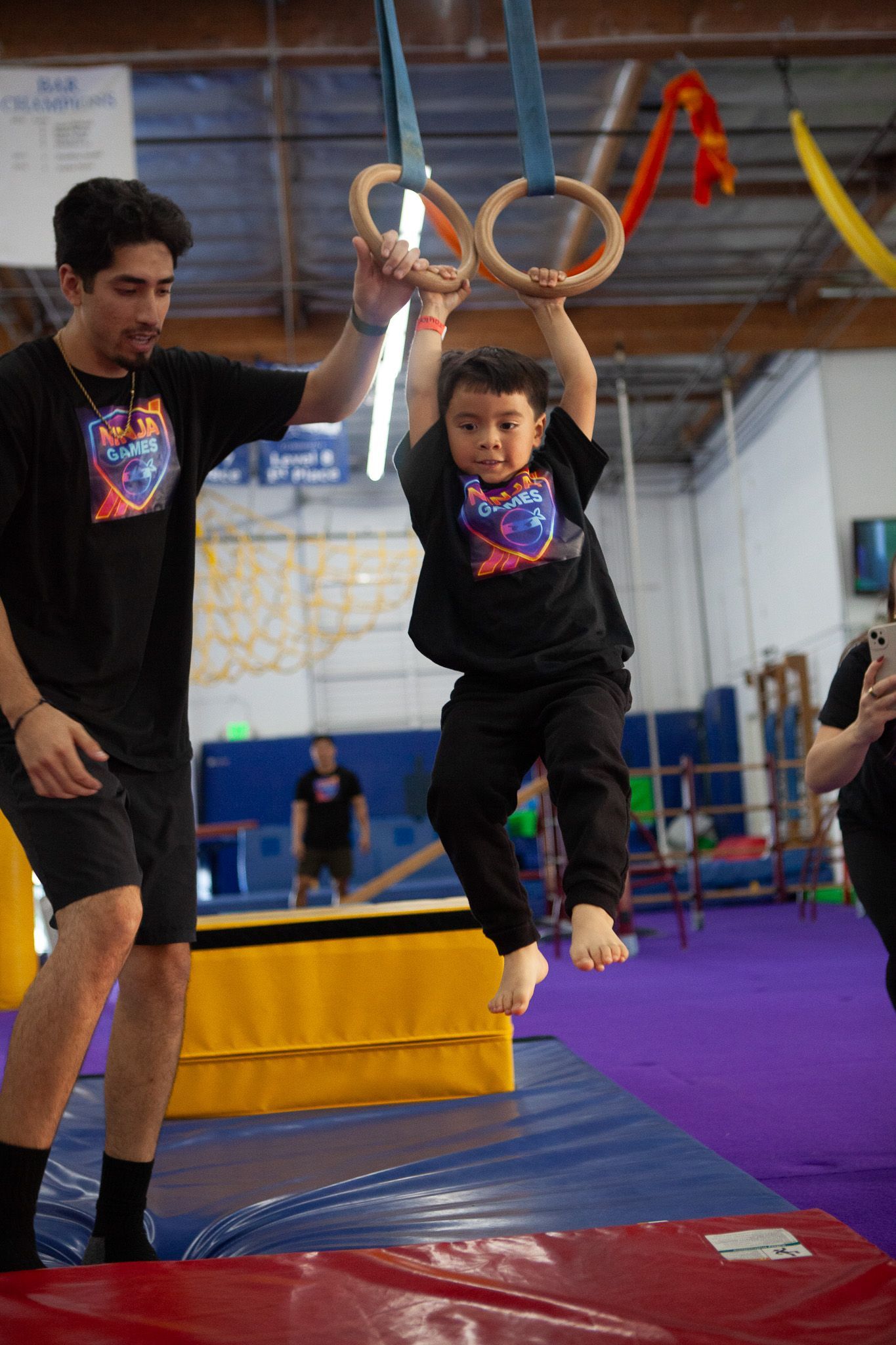 A young child hangs from rings with assistance from an adult in a gym.