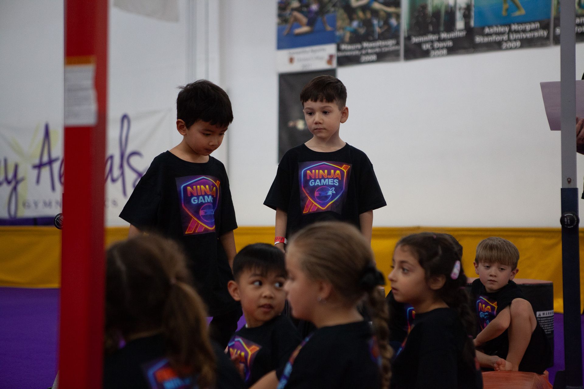 Children in black shirts stand in a gymnastics gym. Some are looking toward the audience while others look at each other.