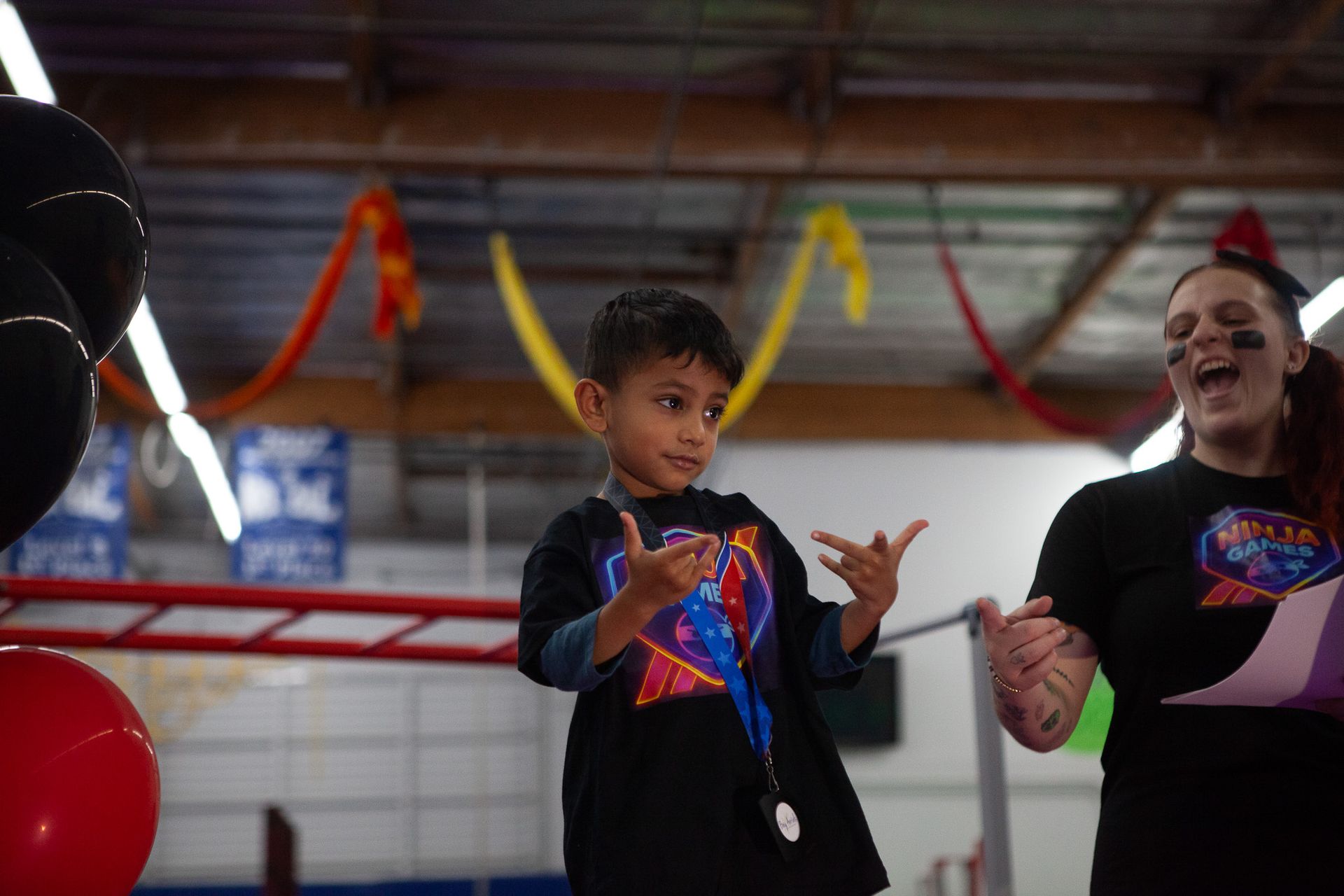 Boy in superhero shirt gestures enthusiastically next to woman holding paper, both indoors, likely at an event.