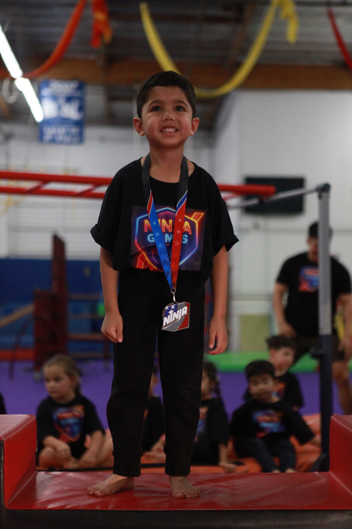Boy standing on a red mat, smiling, in a gym. He is wearing black clothes and a lanyard.