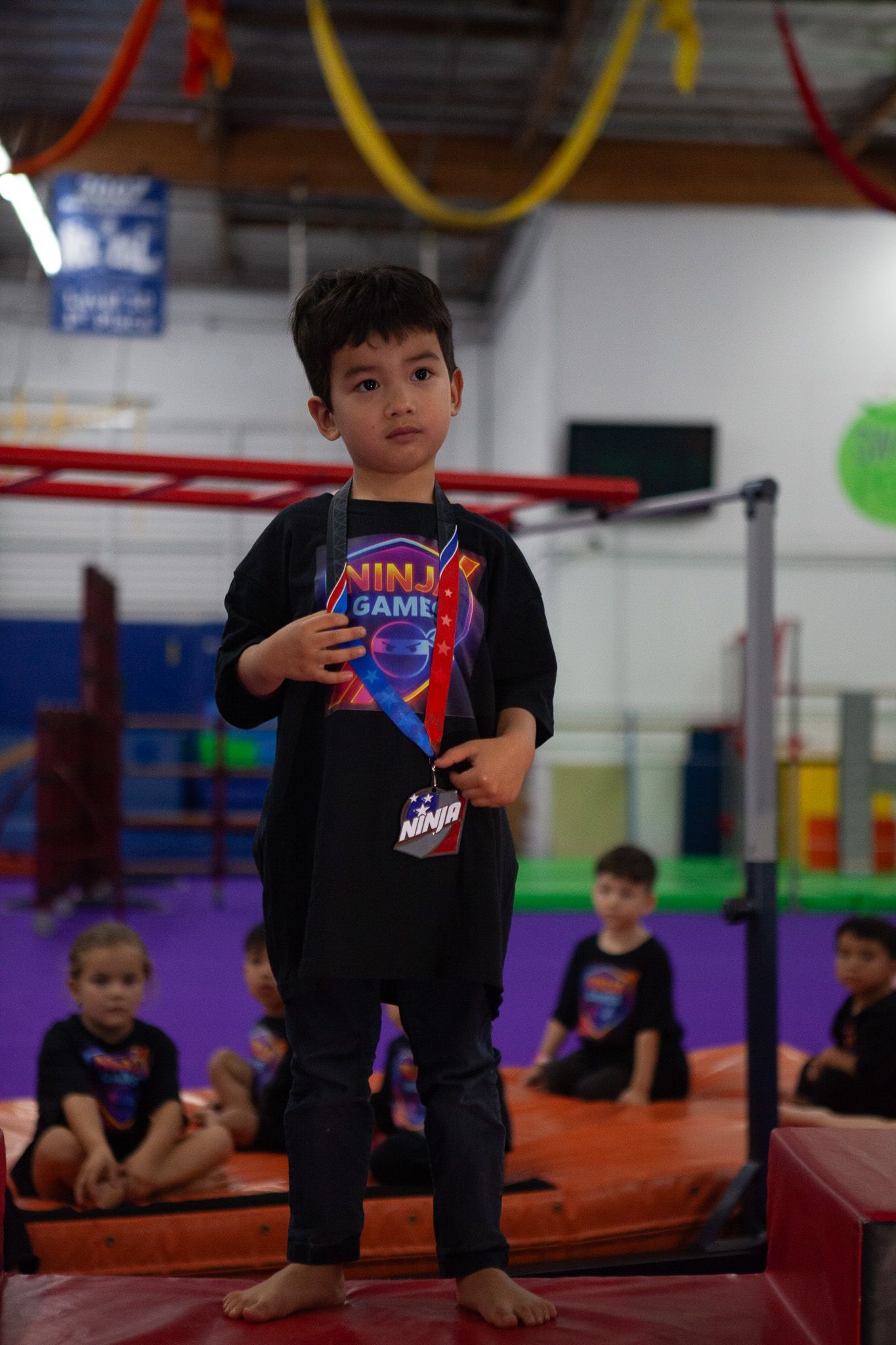 Boy in a black t-shirt and jeans stands with a medal in a gym. Other children are seated on the floor.