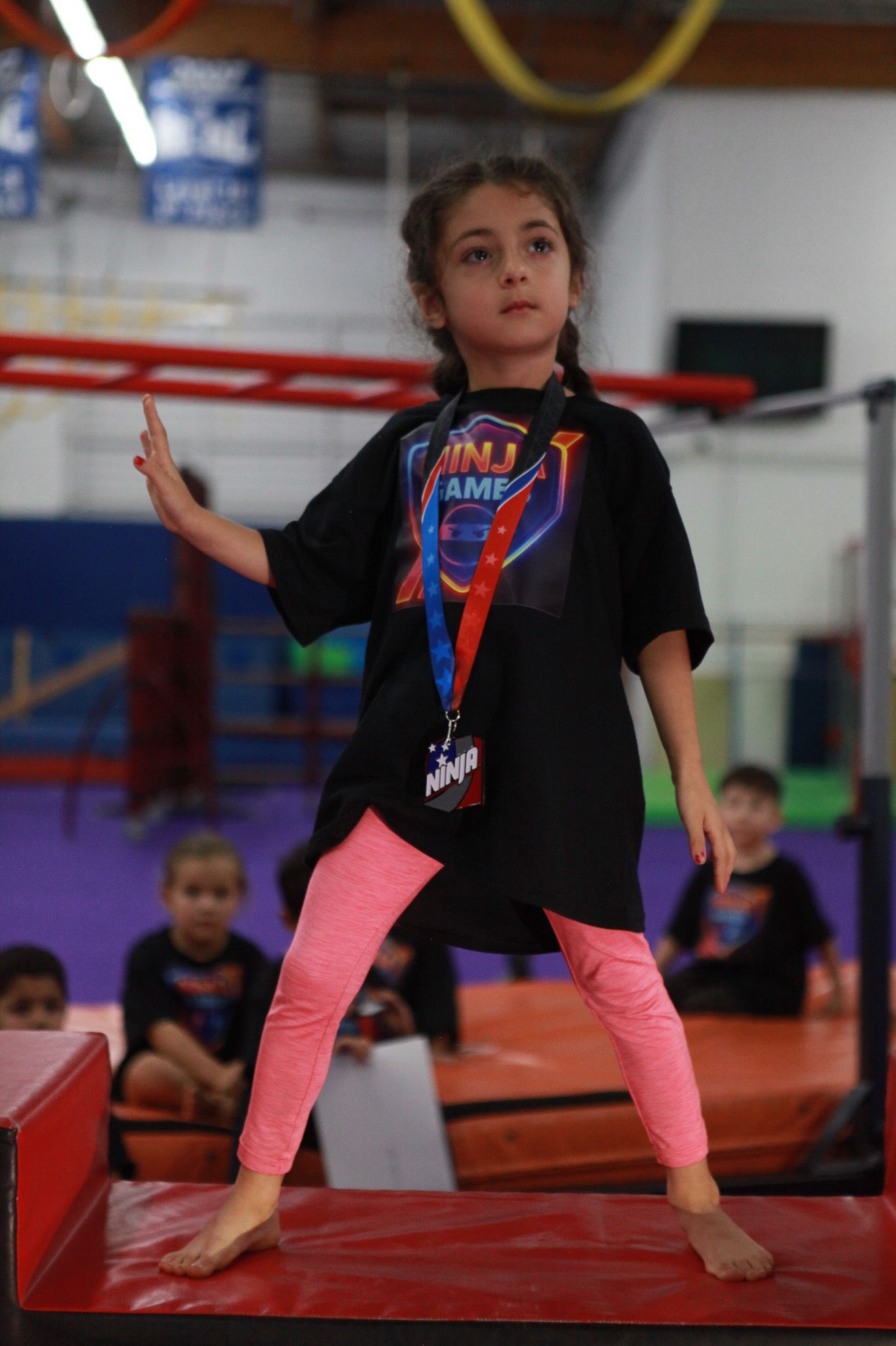 Young girl in pink leggings and black shirt stands with a raised hand, looking toward the side, in a gym.