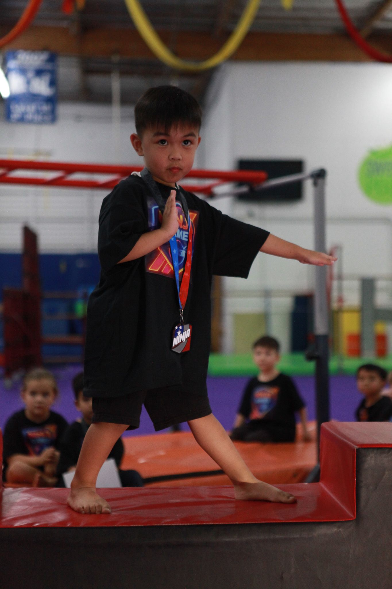 Boy in black shirt and shorts, practicing martial arts stance on a block in a gym.