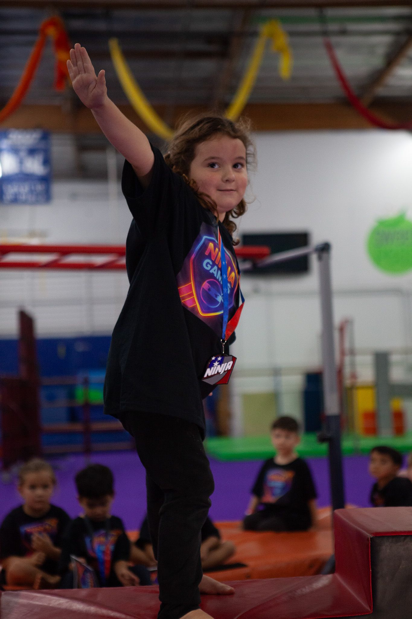 Girl balancing on beam with arm raised, smiling in a gym. Other children watch.