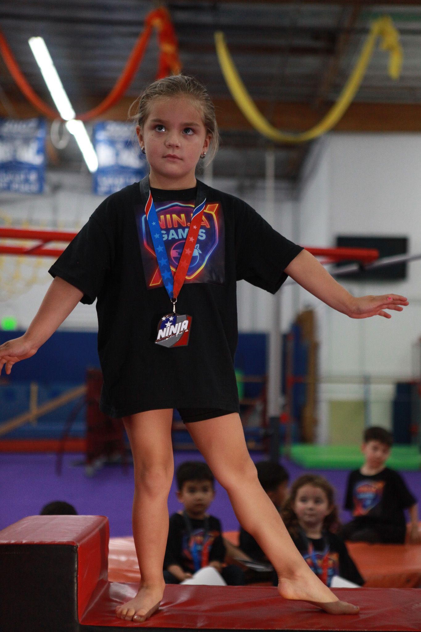 Girl balances on a beam in a gym, arms outstretched, wearing a black shirt and a medal.