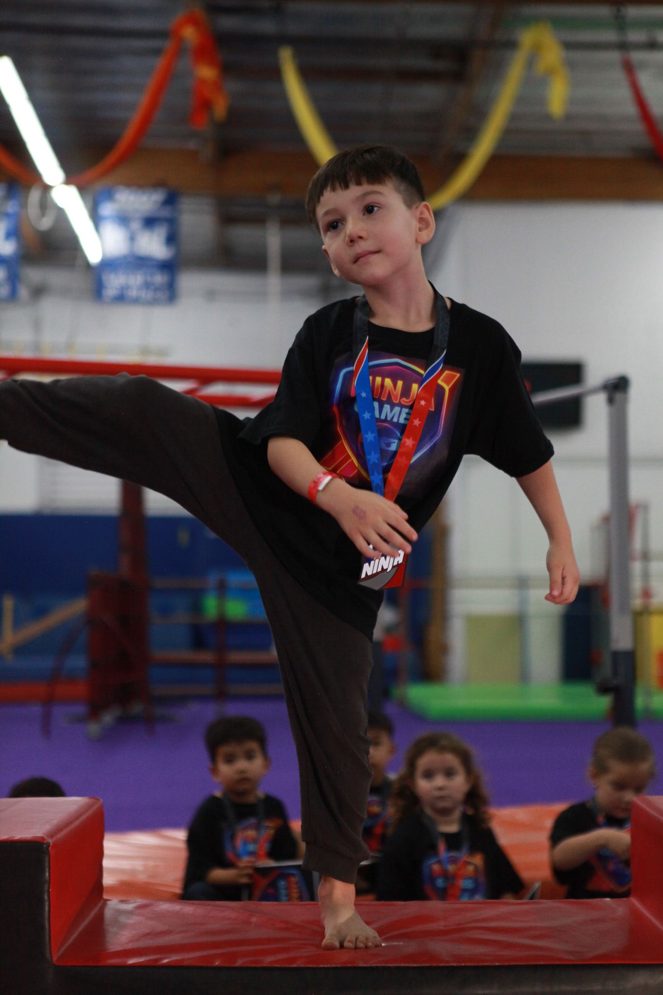 Boy in black shirt and pants kicks; gym setting, other kids watch.