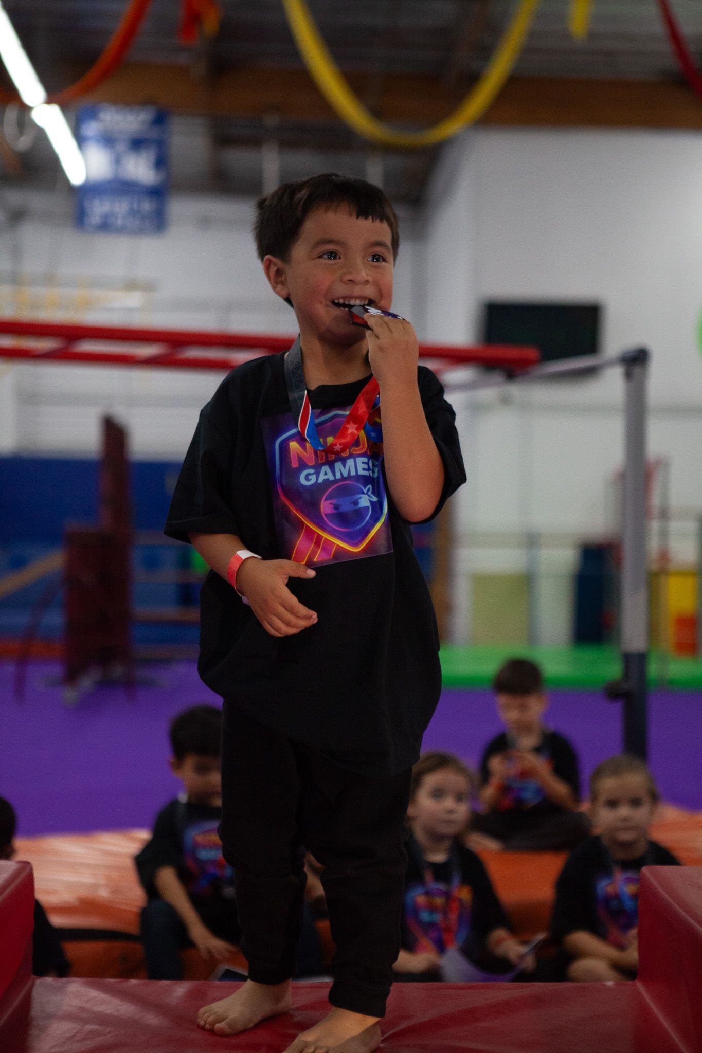 Boy in black shirt and pants with medal; stands on mat in gym. Other kids watch.