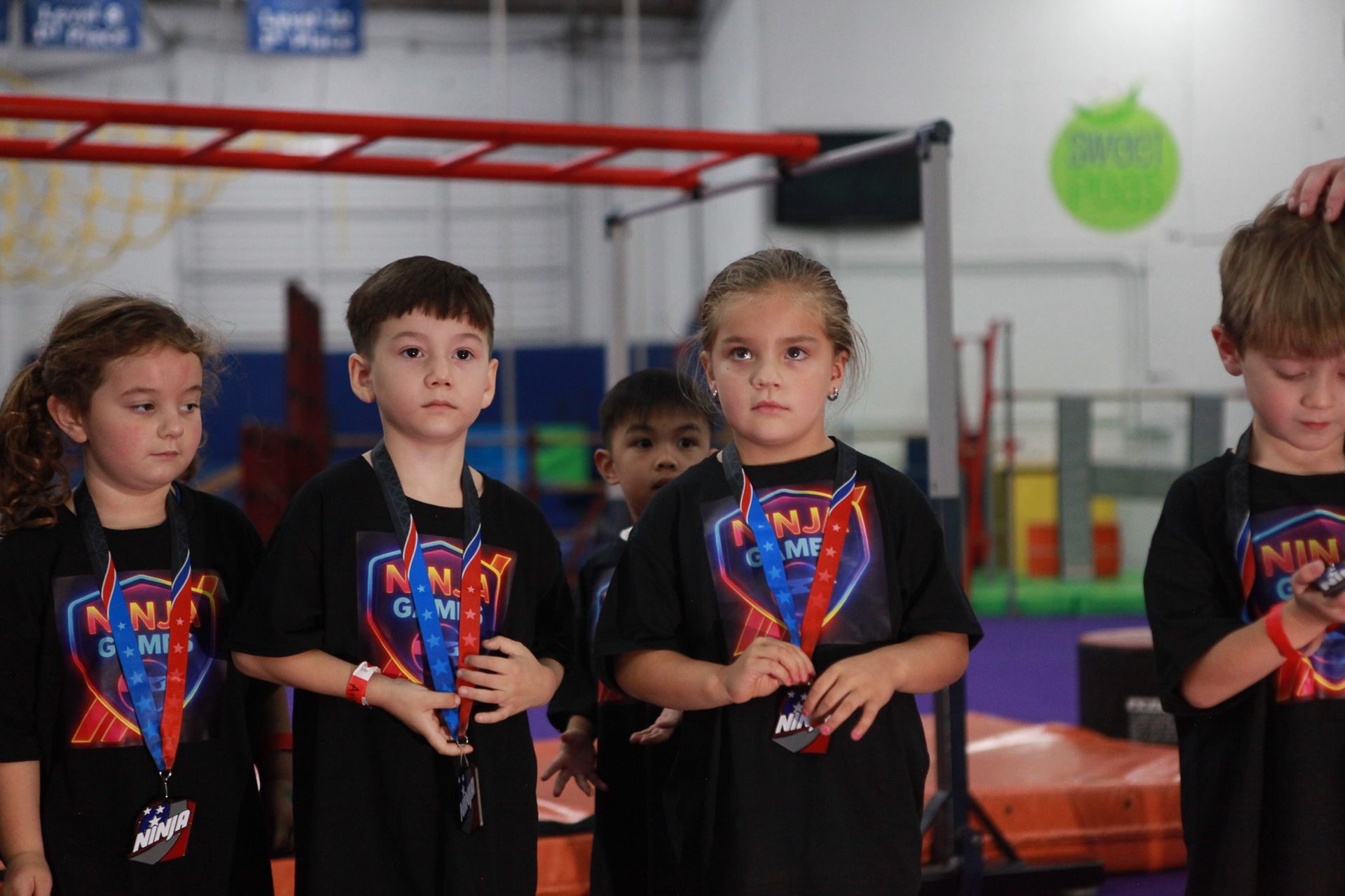 Children with medals stand on a platform; indoor setting. They wear matching black shirts with logos, looking forward.