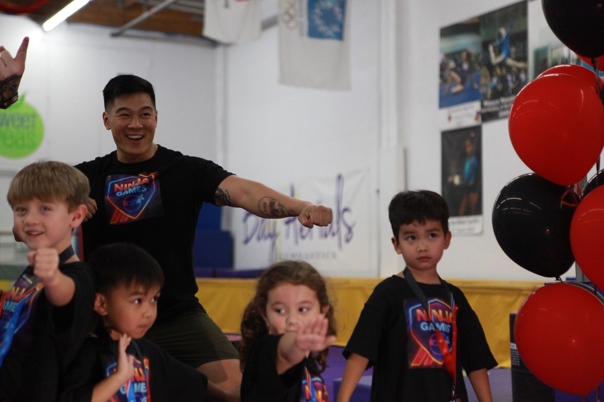 Man and four children smiling, posing in a gym near red and black balloons. They wear matching black shirts.