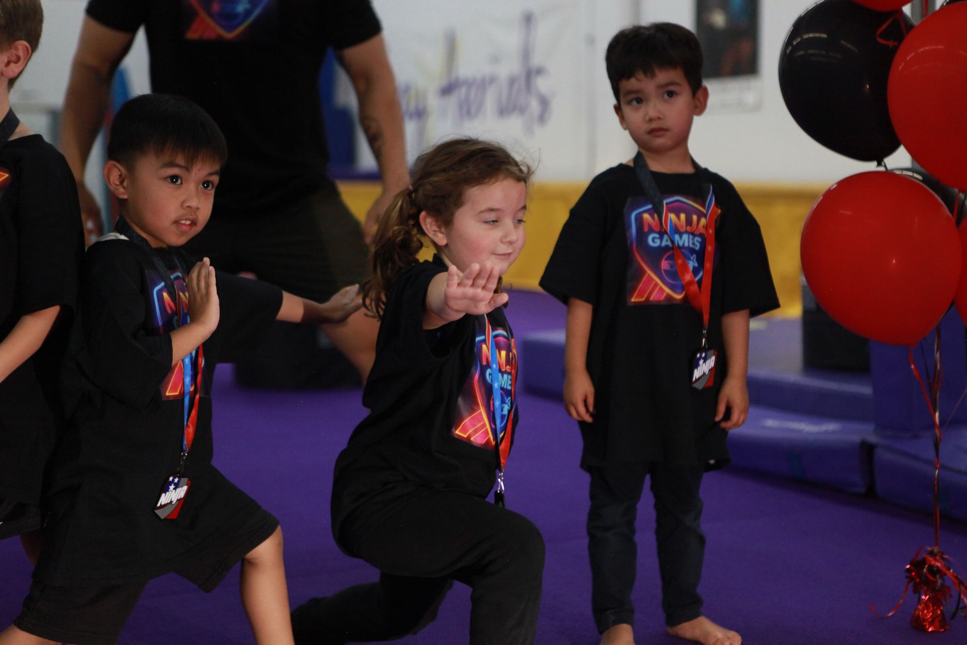 Children in black shirts practicing martial arts on a purple mat, balloons in the background.
