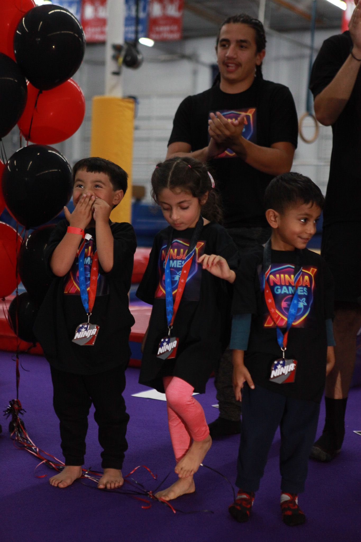 Children at a gymnastics event stand, watching, with balloons. A man in black shirt claps in the background.