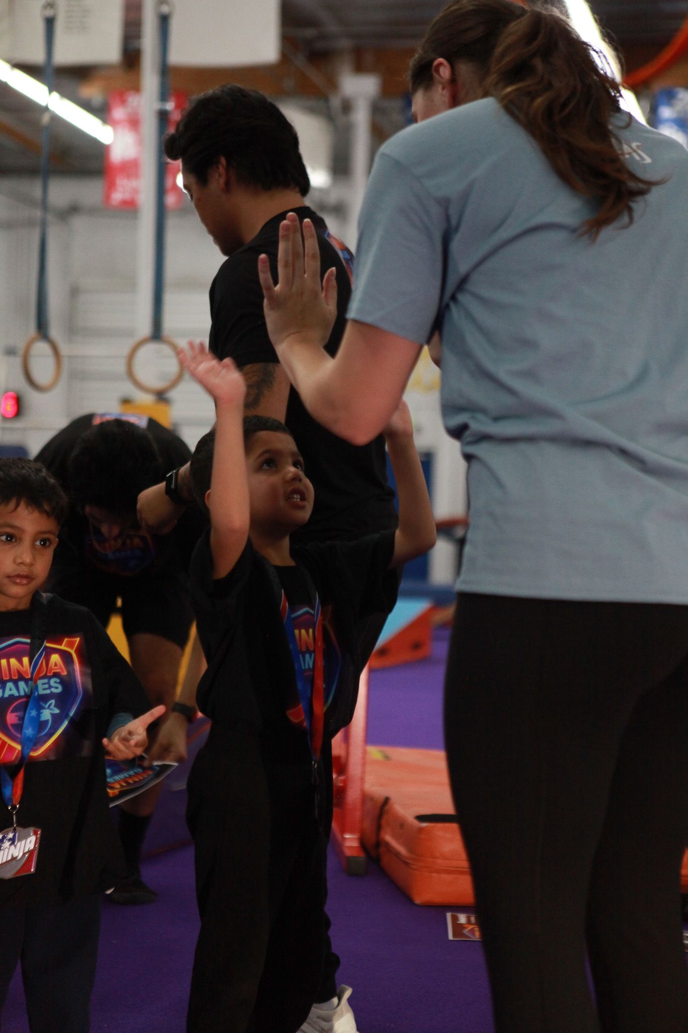 Young child with raised arm high-fiving a person in a gym. Other people and equipment in the background.