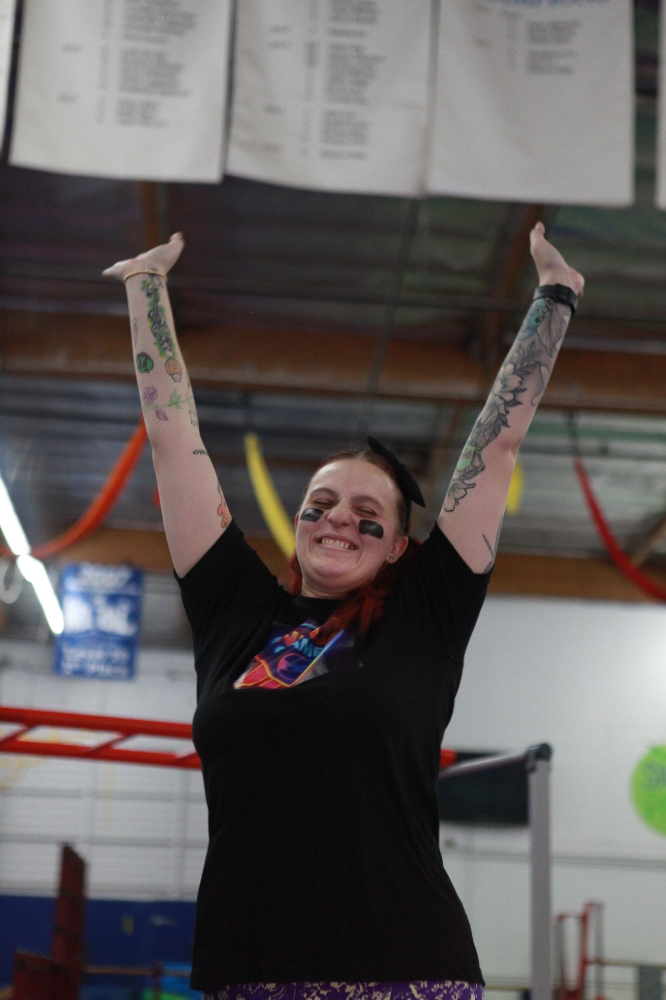 Person with arm tattoos raises arms in celebration, wearing a black shirt and face paint indoors.