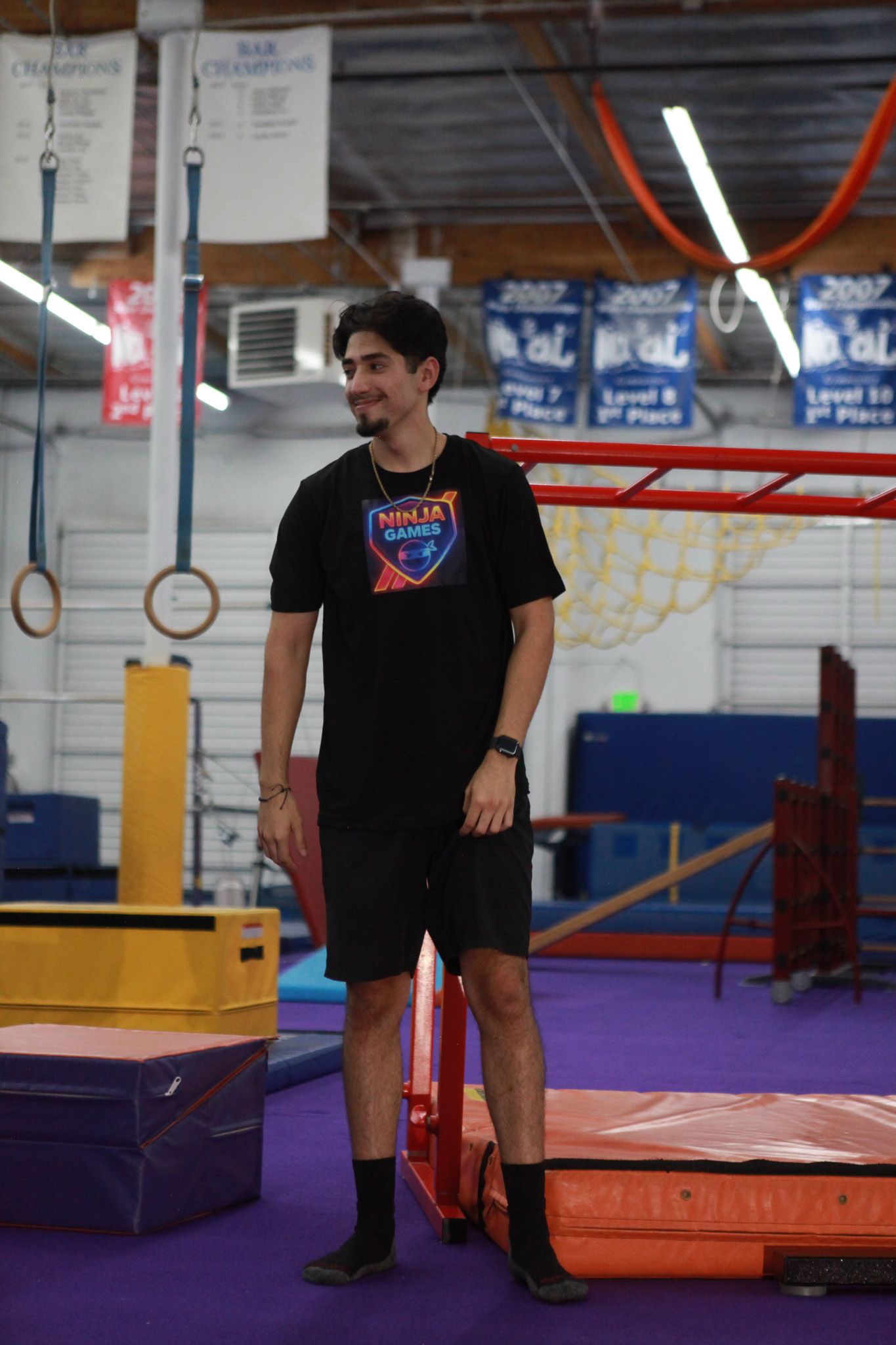 Man in black shirt and shorts smiles in a gym, near obstacle course equipment and mats.