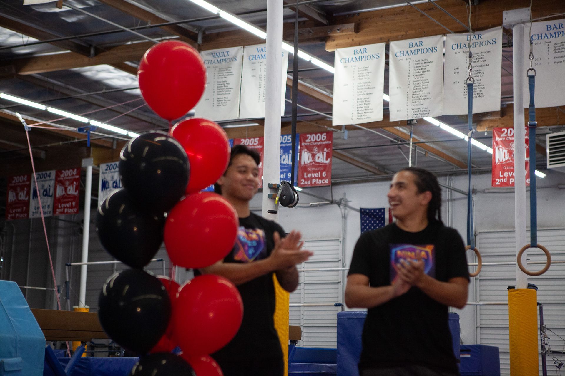 Two men clapping in a gym with balloons; red and black. Banners hang in the background.