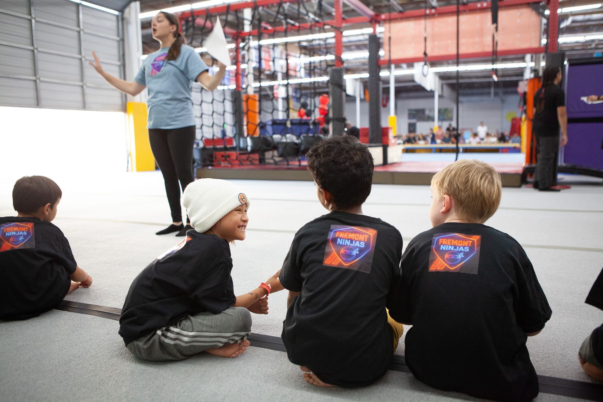 Children in black shirts listen to instructor in gym, with obstacles visible.