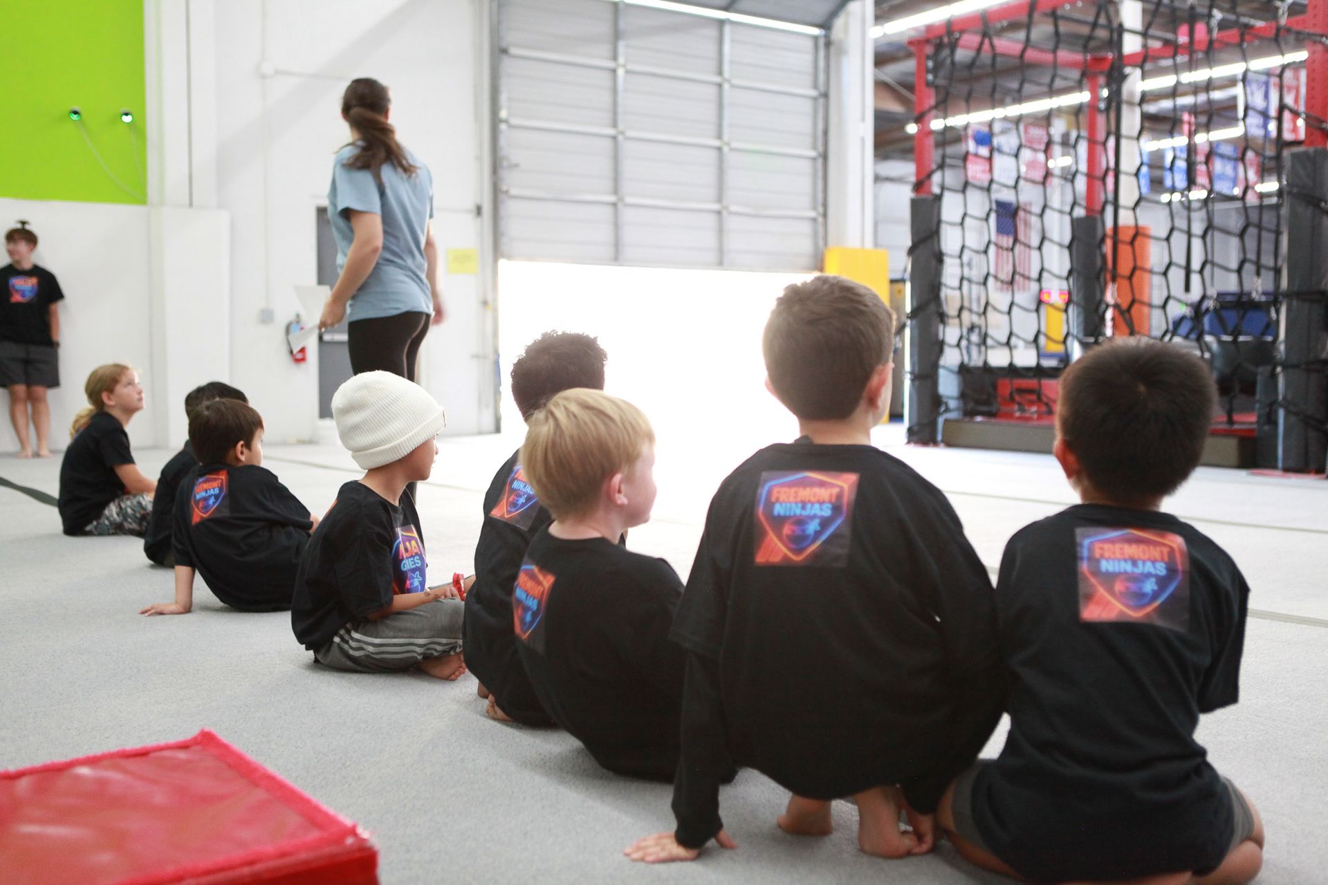 Children in black shirts, kneeling, watch an instructor at an indoor sports facility.