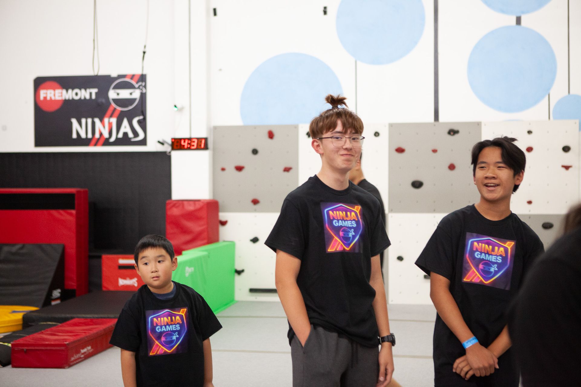 Four people in black shirts at a ninja gym. The middle person has a bun, and all are smiling. 