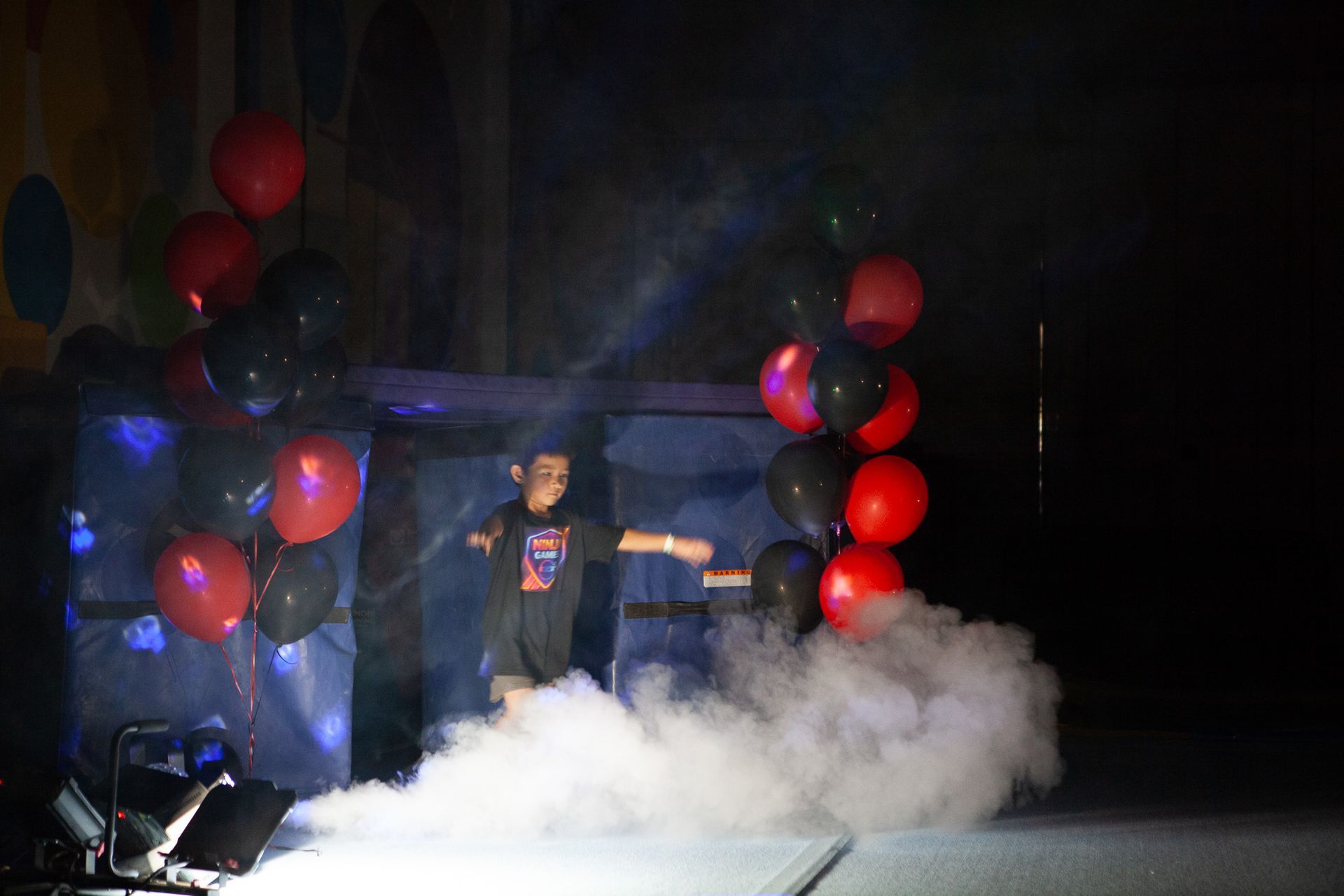 A person on stage with smoke and balloons. Red and black balloons arch over them, smoke billows at their feet.