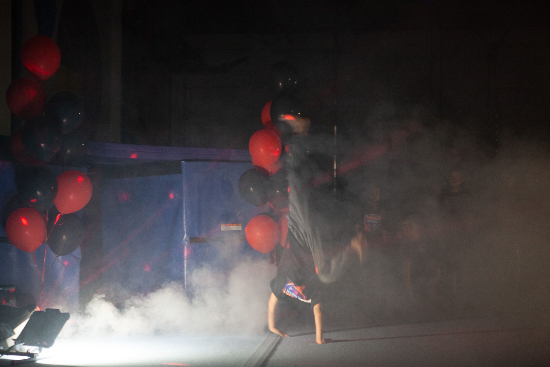 Dancer doing a handstand on stage with red and black balloons, smoke, and blue lights in the background.