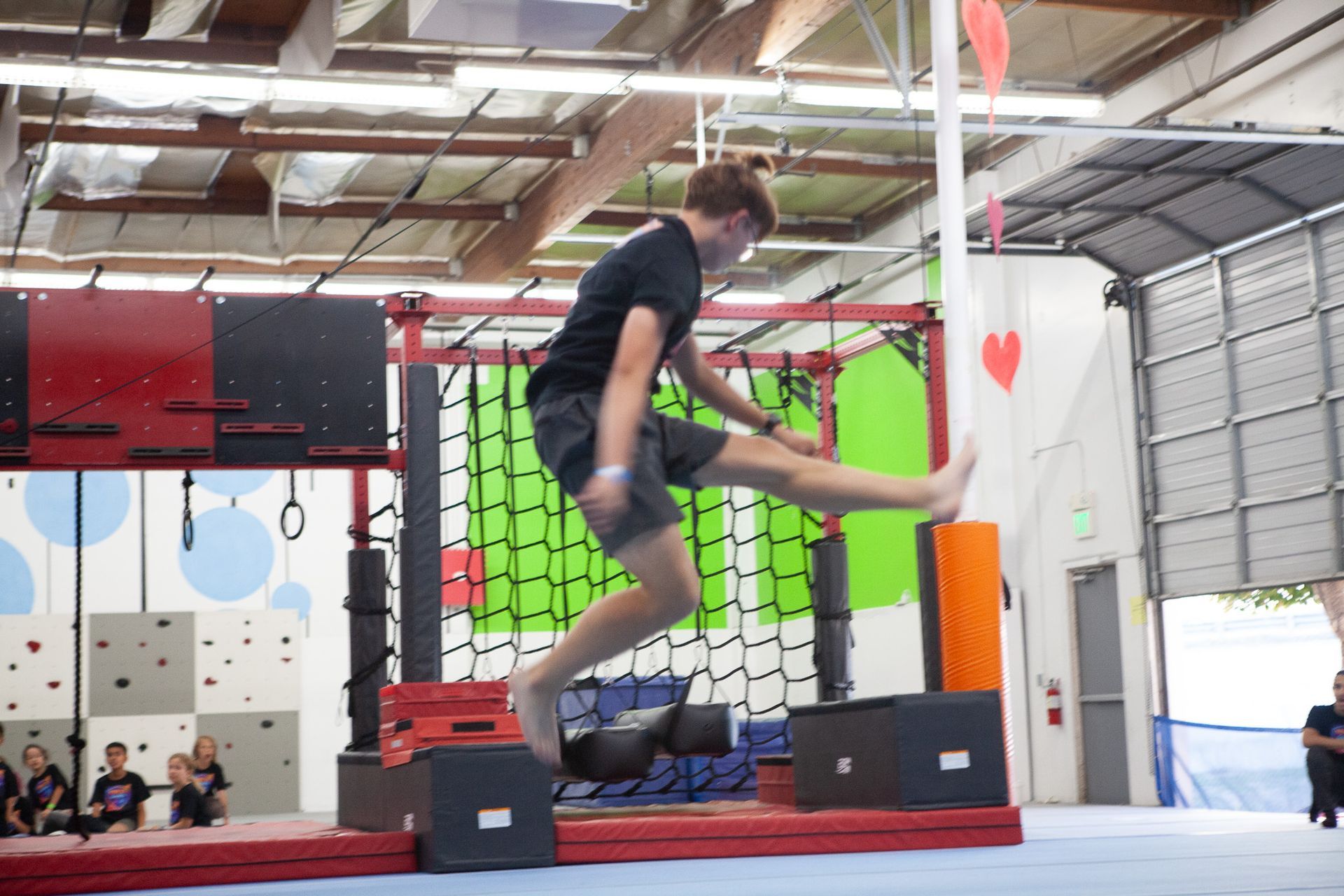 Teenager jumping over obstacles in an indoor obstacle course; black shirt, gray shorts.