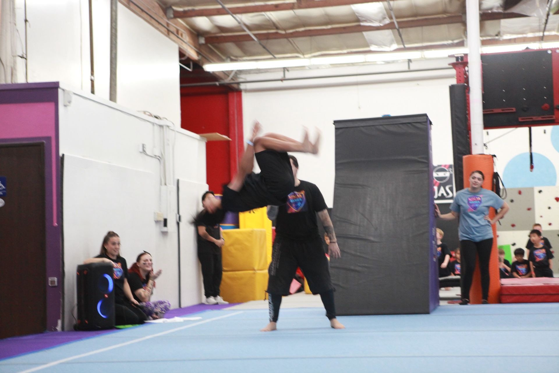 Man doing backflip over another man in an indoor gym setting. People watching.