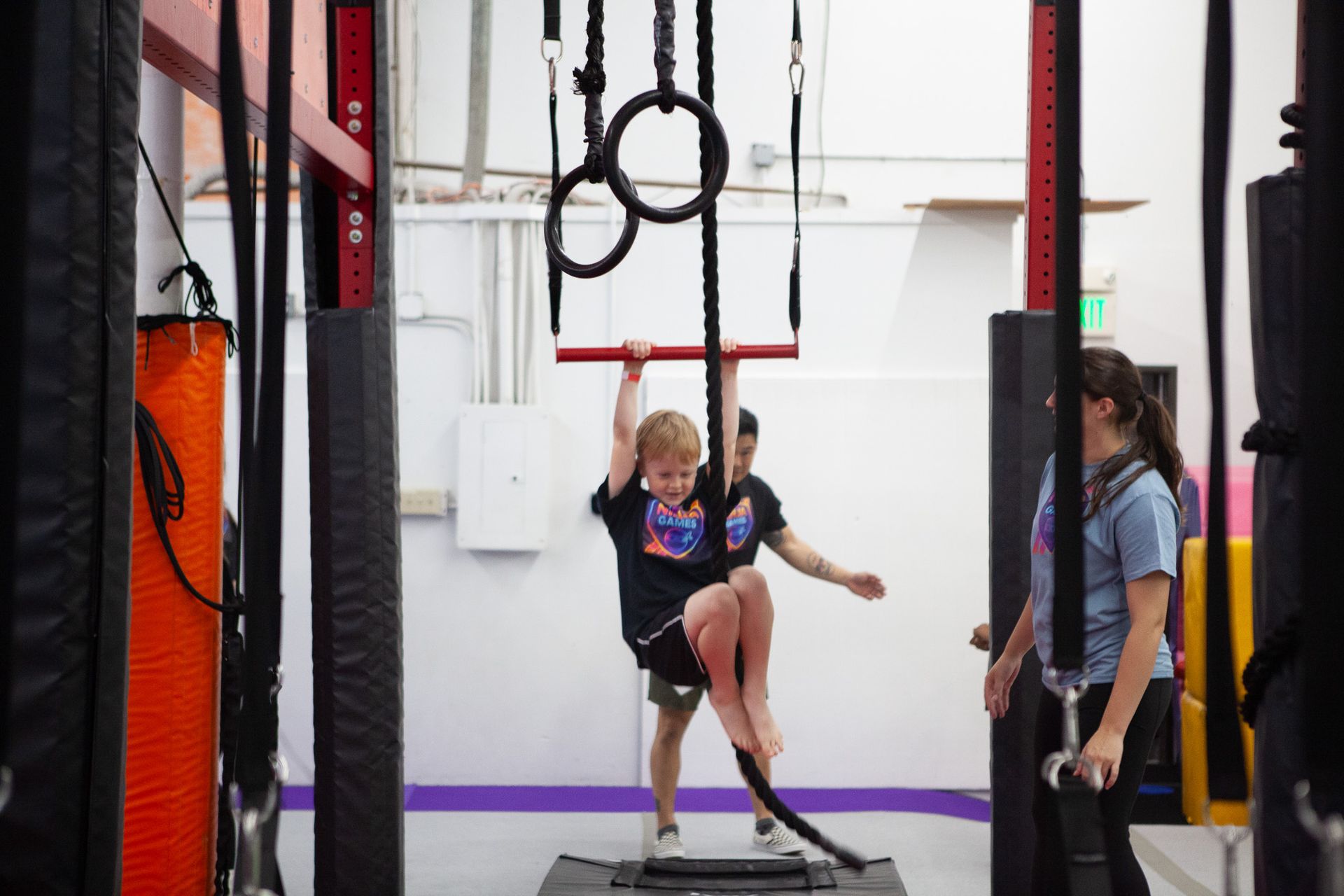 Boy climbing a rope in an indoor obstacle course, assisted by two adults.