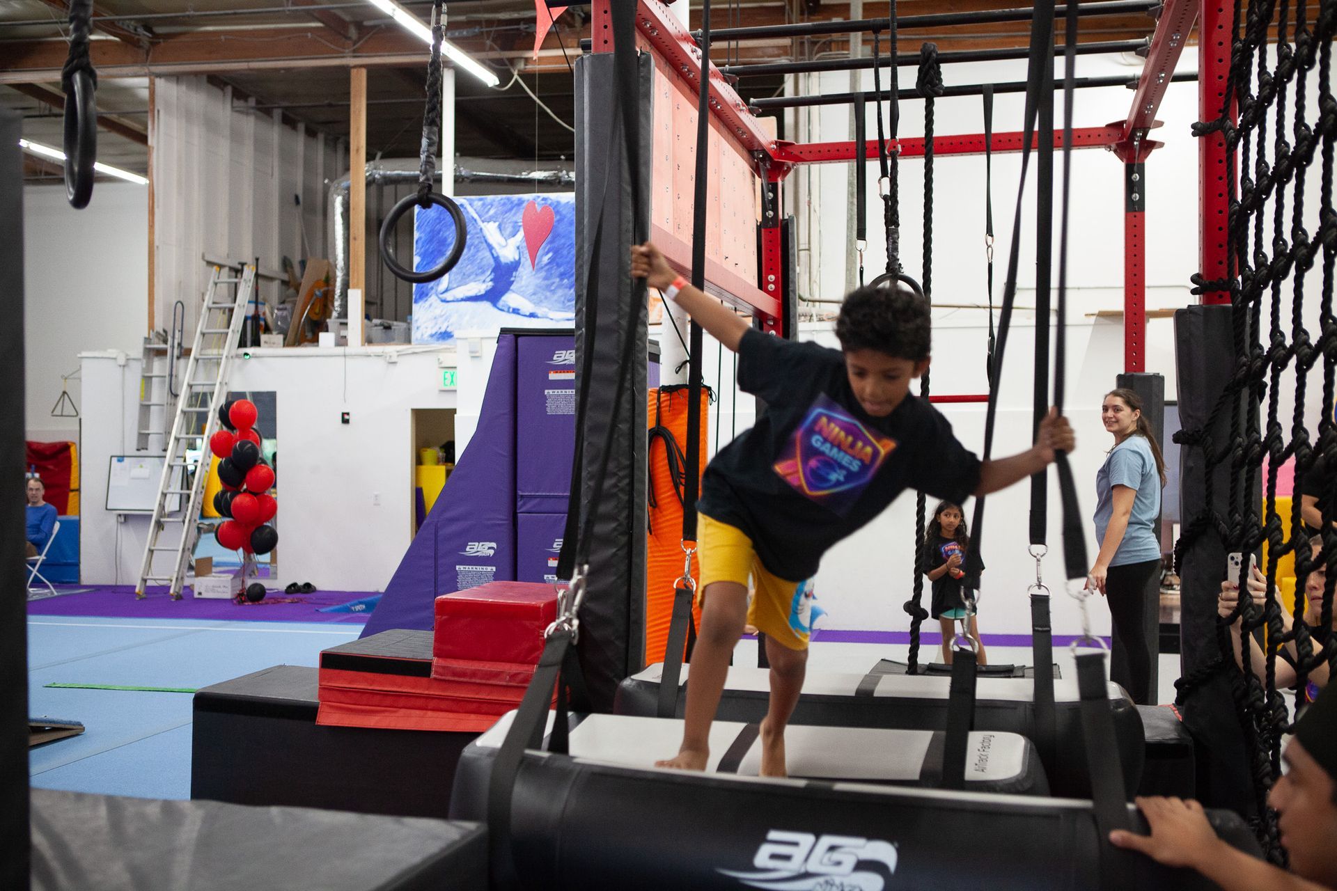 Boy on obstacle course, grasping hanging ropes and traversing a platform in a gym, wearing a t-shirt and yellow shorts.