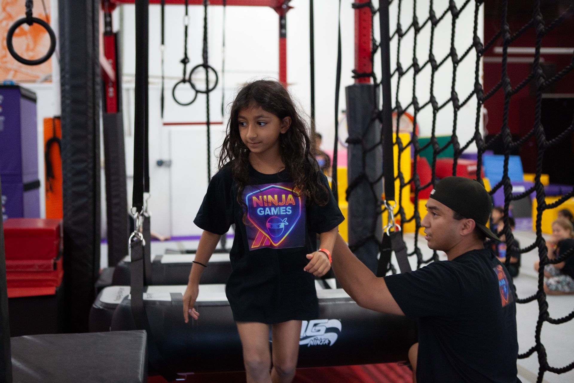 A girl in a black shirt walks a ninja course with an instructor's assistance.