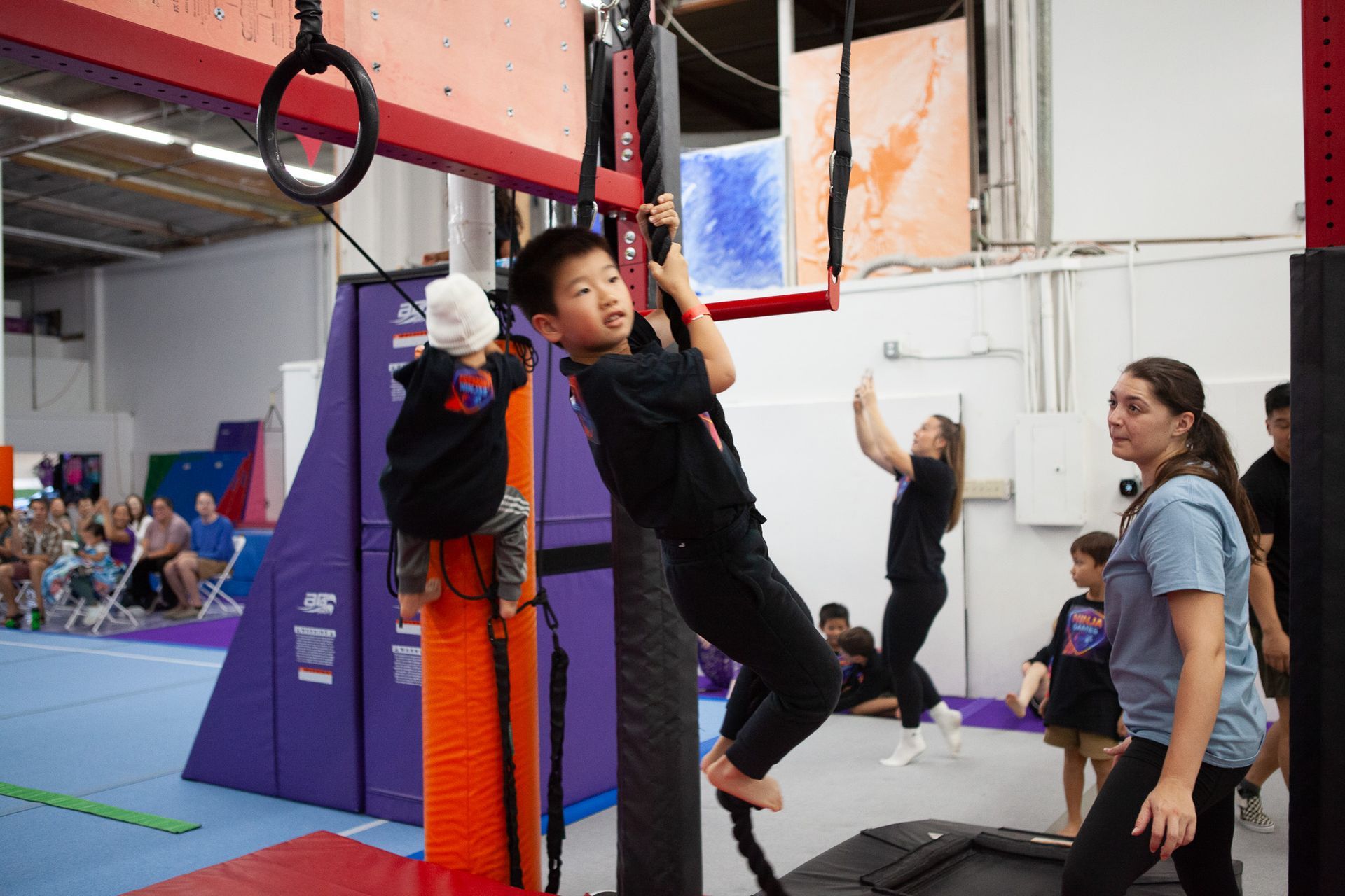 Boy hanging from rope on obstacle course, other children and adult in background.