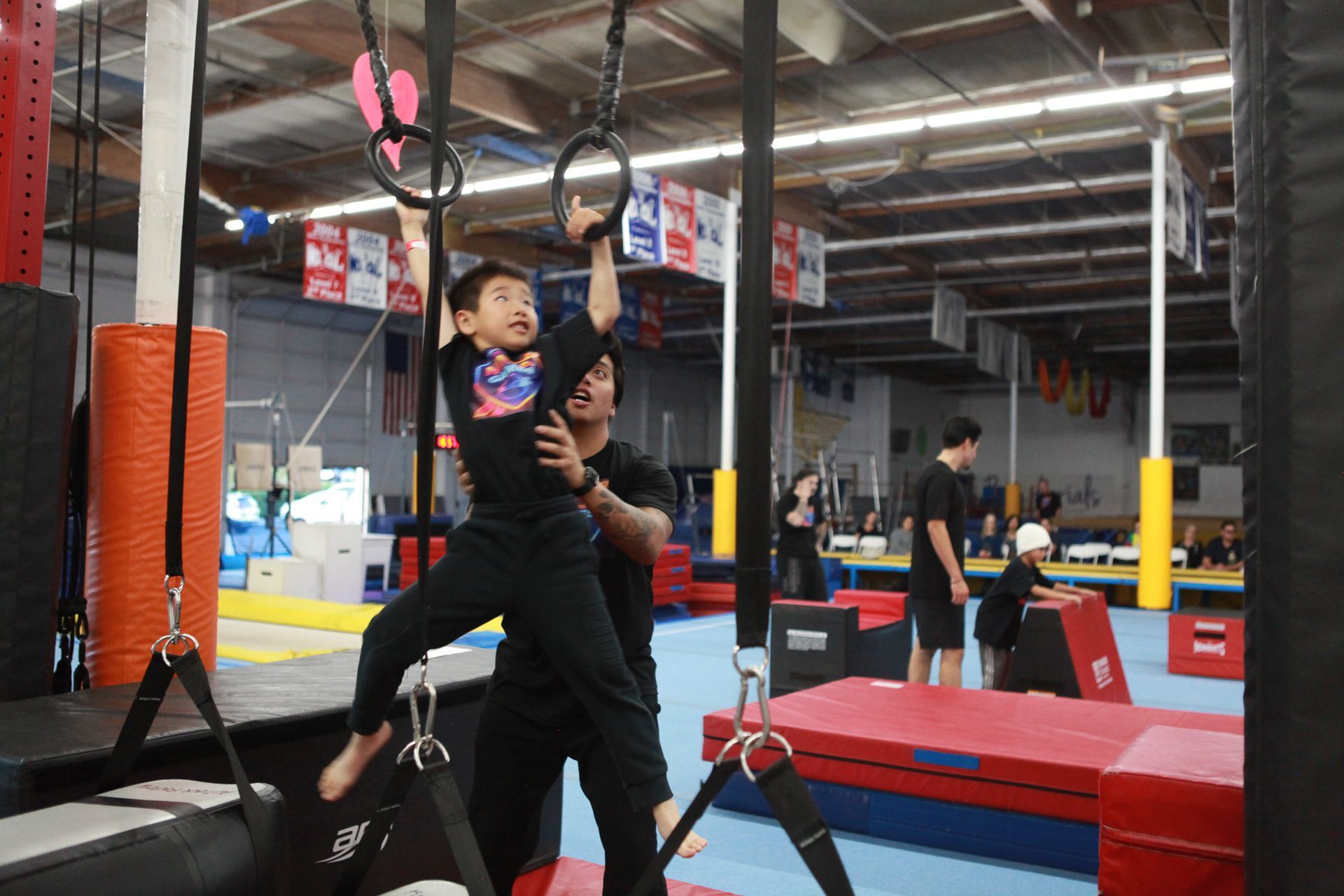 Boy hanging from rings, assisted by an adult in an obstacle gym; orange, red, and blue mats.