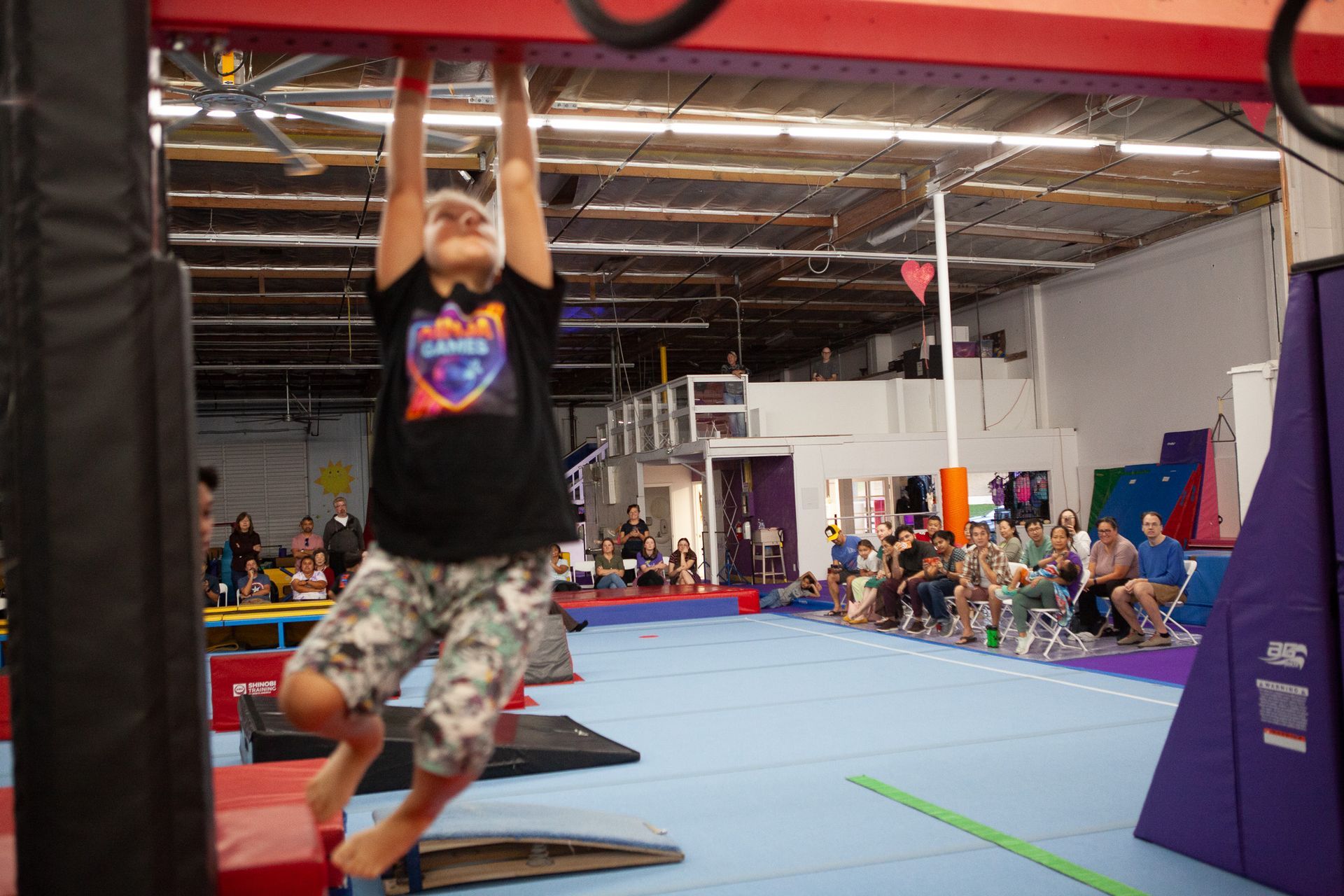 Person hangs from a red beam at an obstacle course gym. People watch in background.