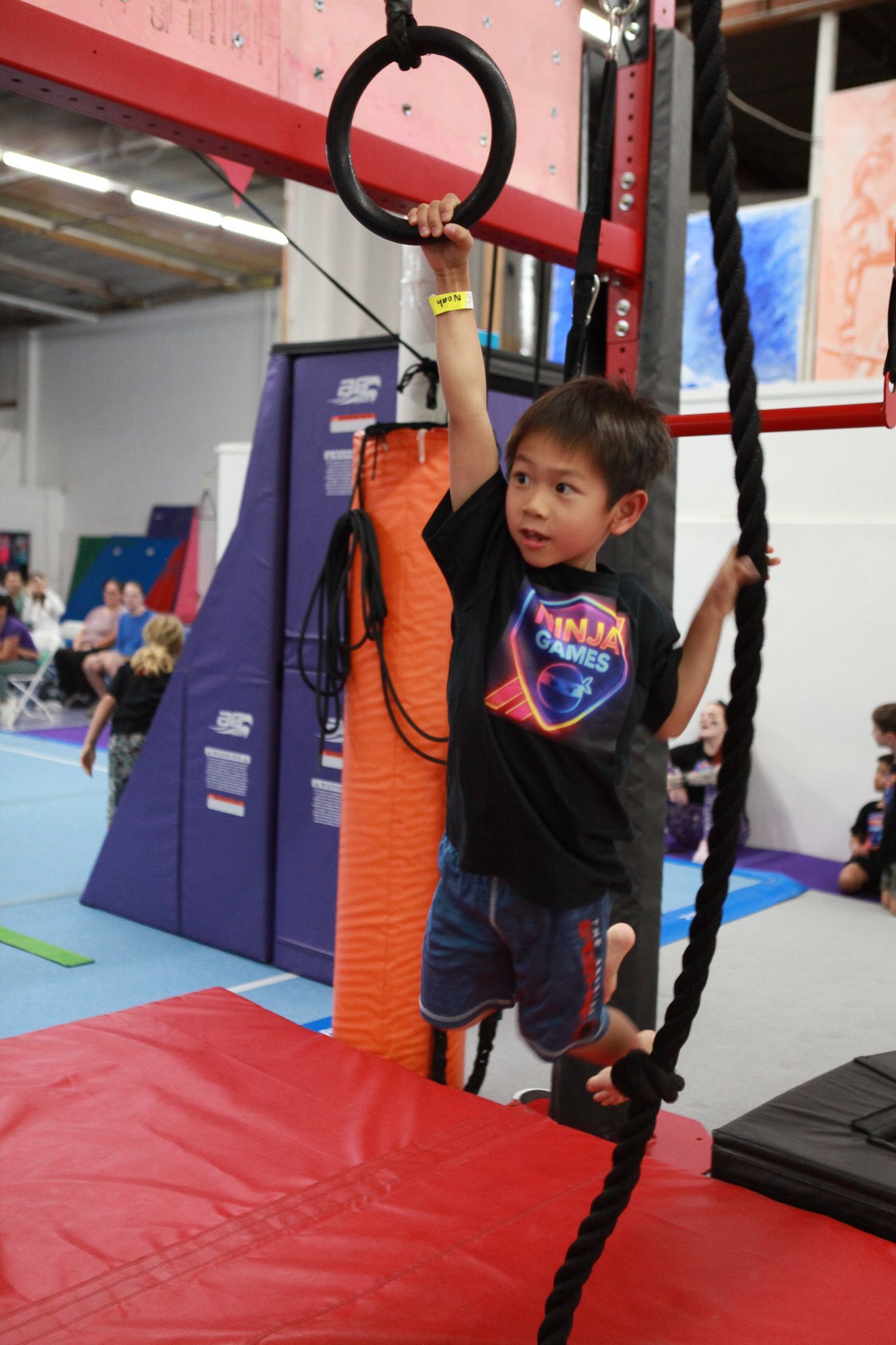 Boy hanging from a ring and rope in a gym. He is wearing a black shirt and blue shorts.