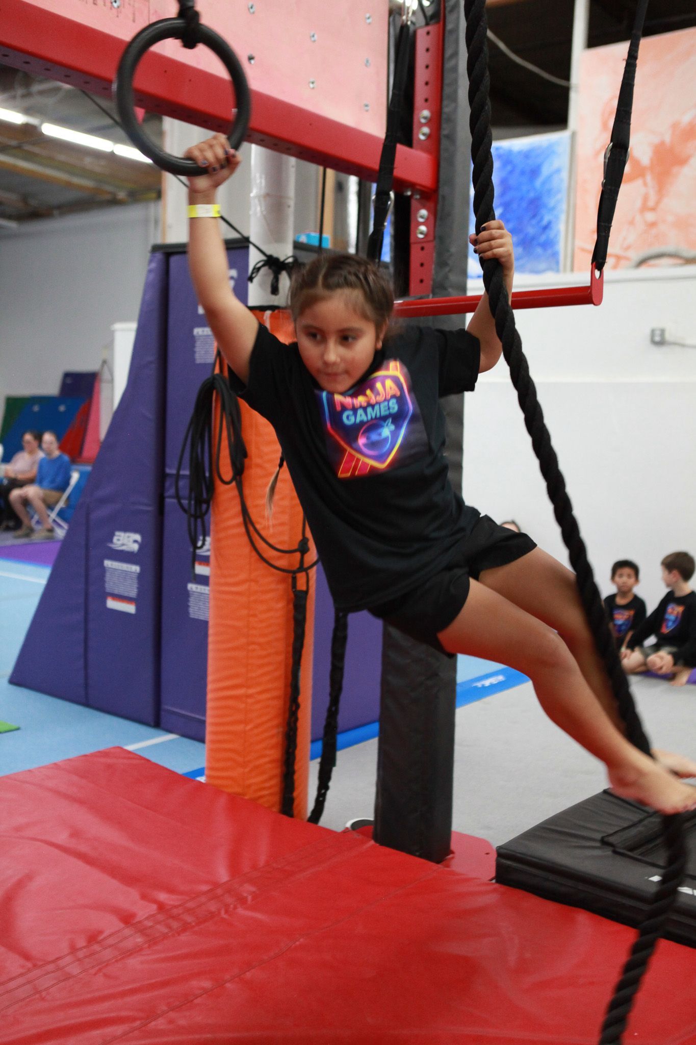 Girl on gymnastics apparatus, holding a ring, rope in the other hand. Indoor gym setting.