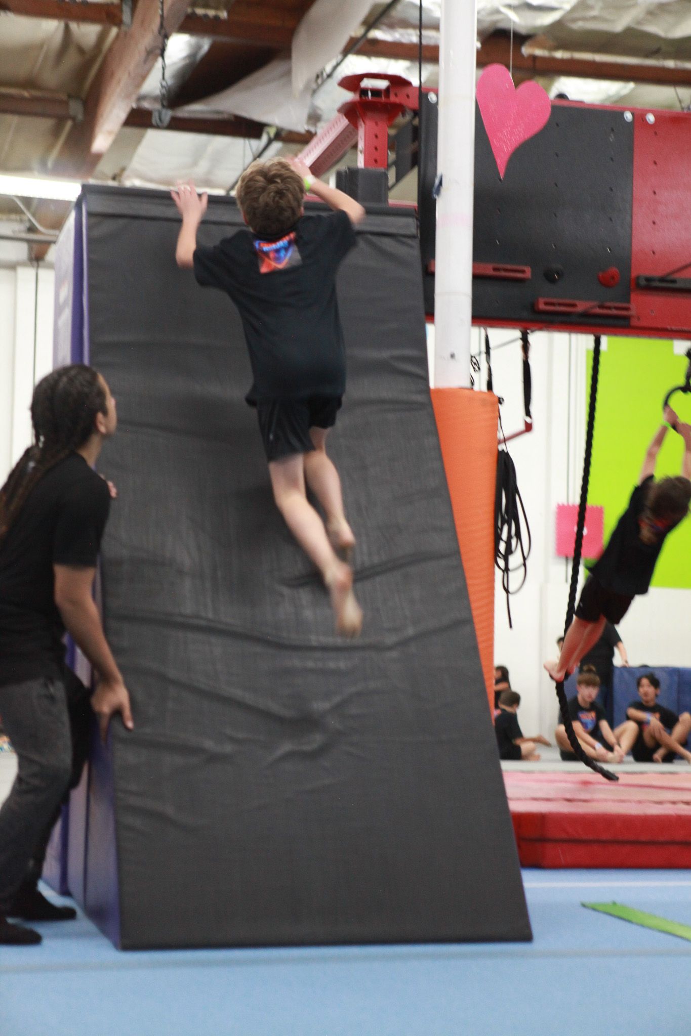 Boy attempting to scale a slanted climbing wall in a gym, with help from a spotter; another person on rings.