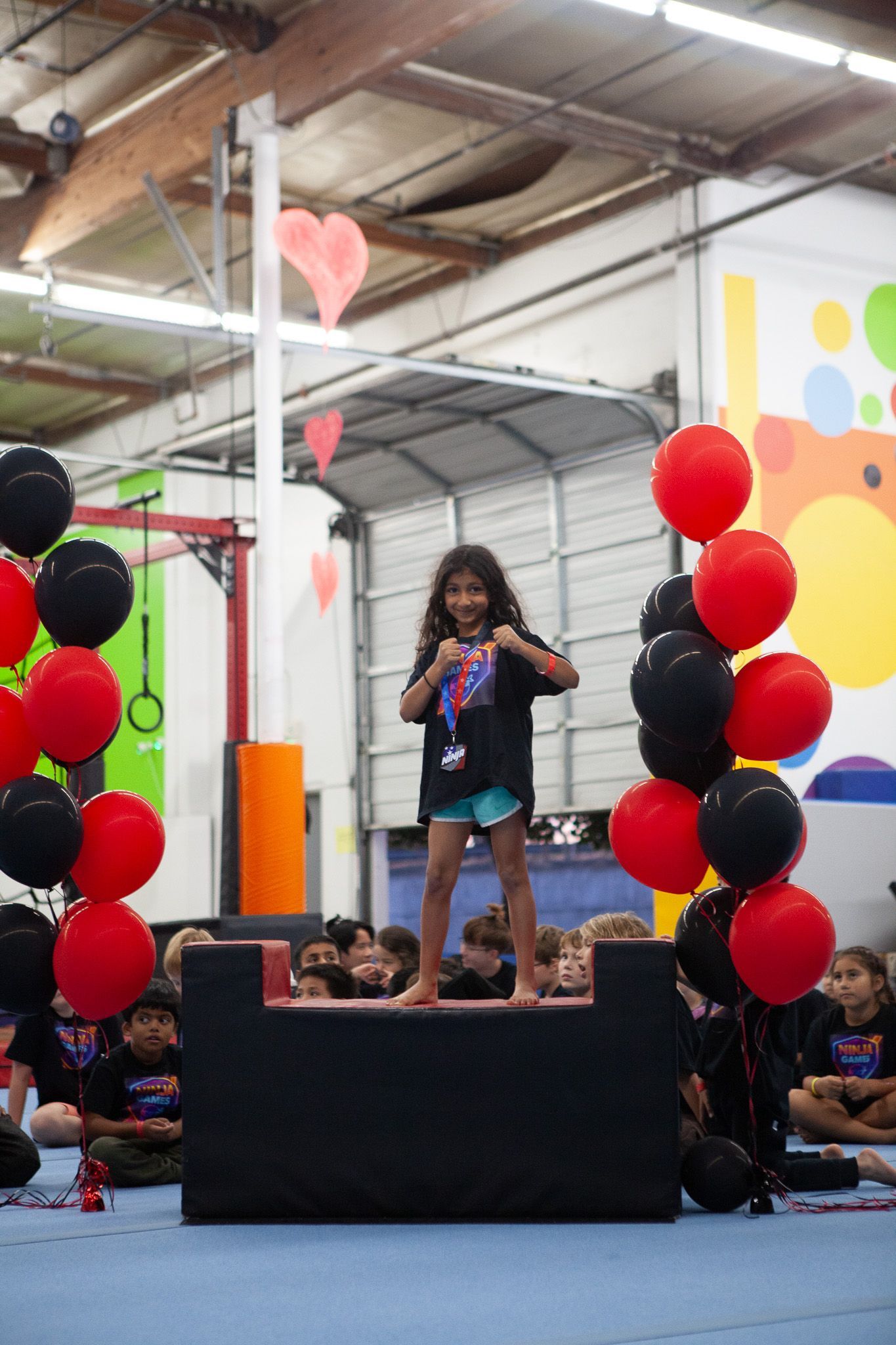 Girl stands on a platform, martial arts pose. Balloons frame her. Gym interior.