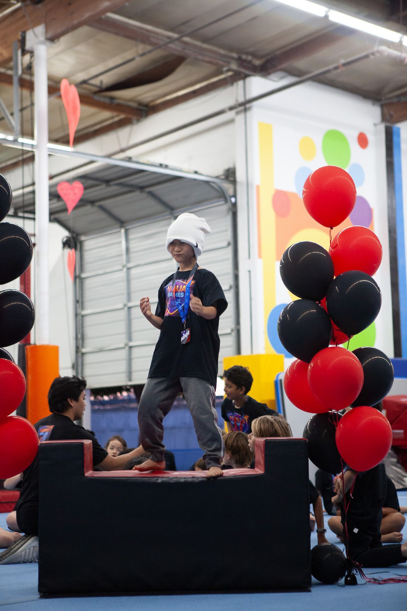 Child in chef's hat standing on black block, surrounded by red and black balloons, inside a gym.