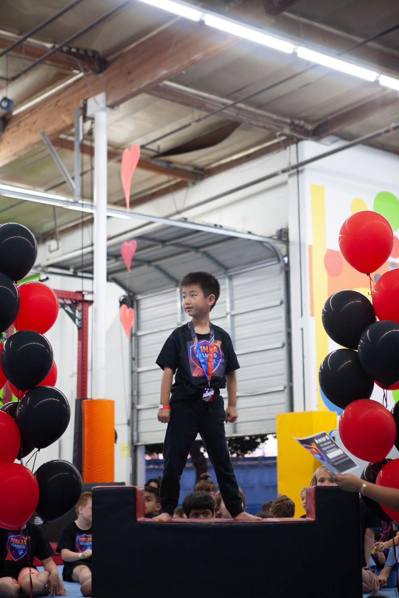 Young child on a stage, smiling, surrounded by red and black balloons, at an indoor event.