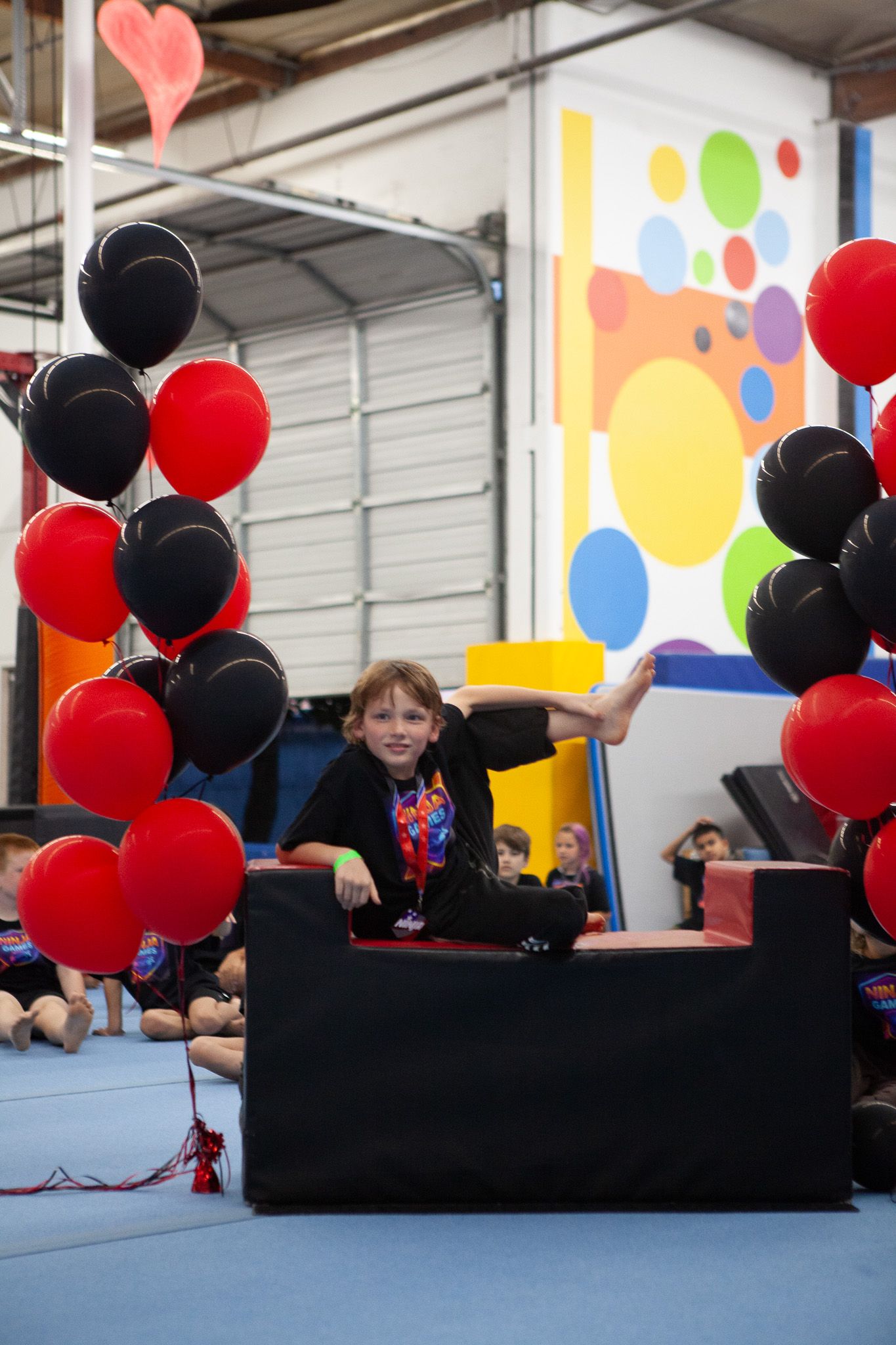 Boy sitting on black foam block at a gym with red and black balloons, smiling.