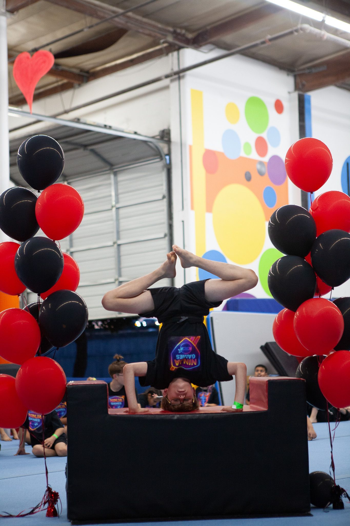 Boy balances upside down on a black block with red and black balloons; gym setting.