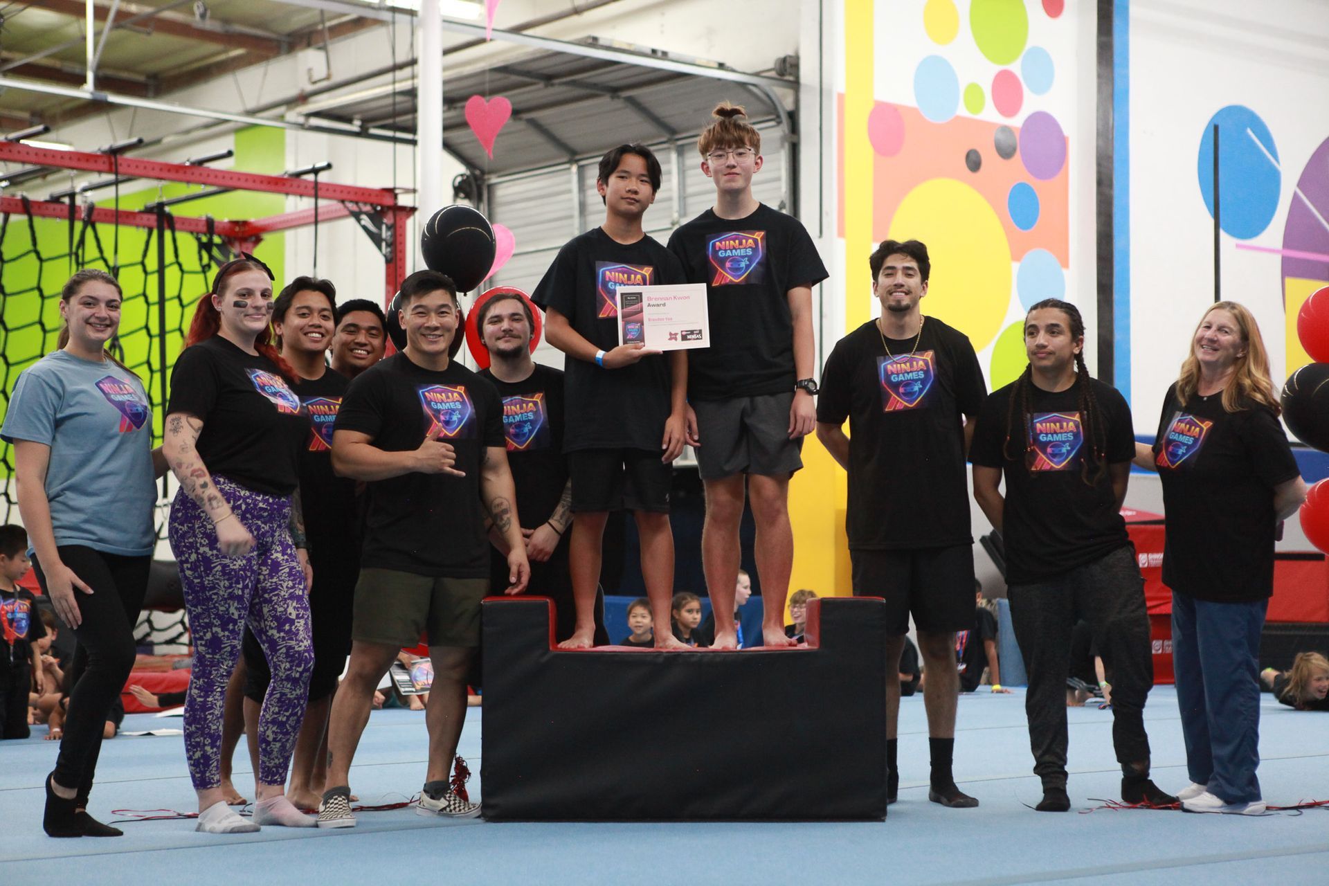 Group poses with a certificate, standing on a podium. They wear matching black shirts in an indoor setting.