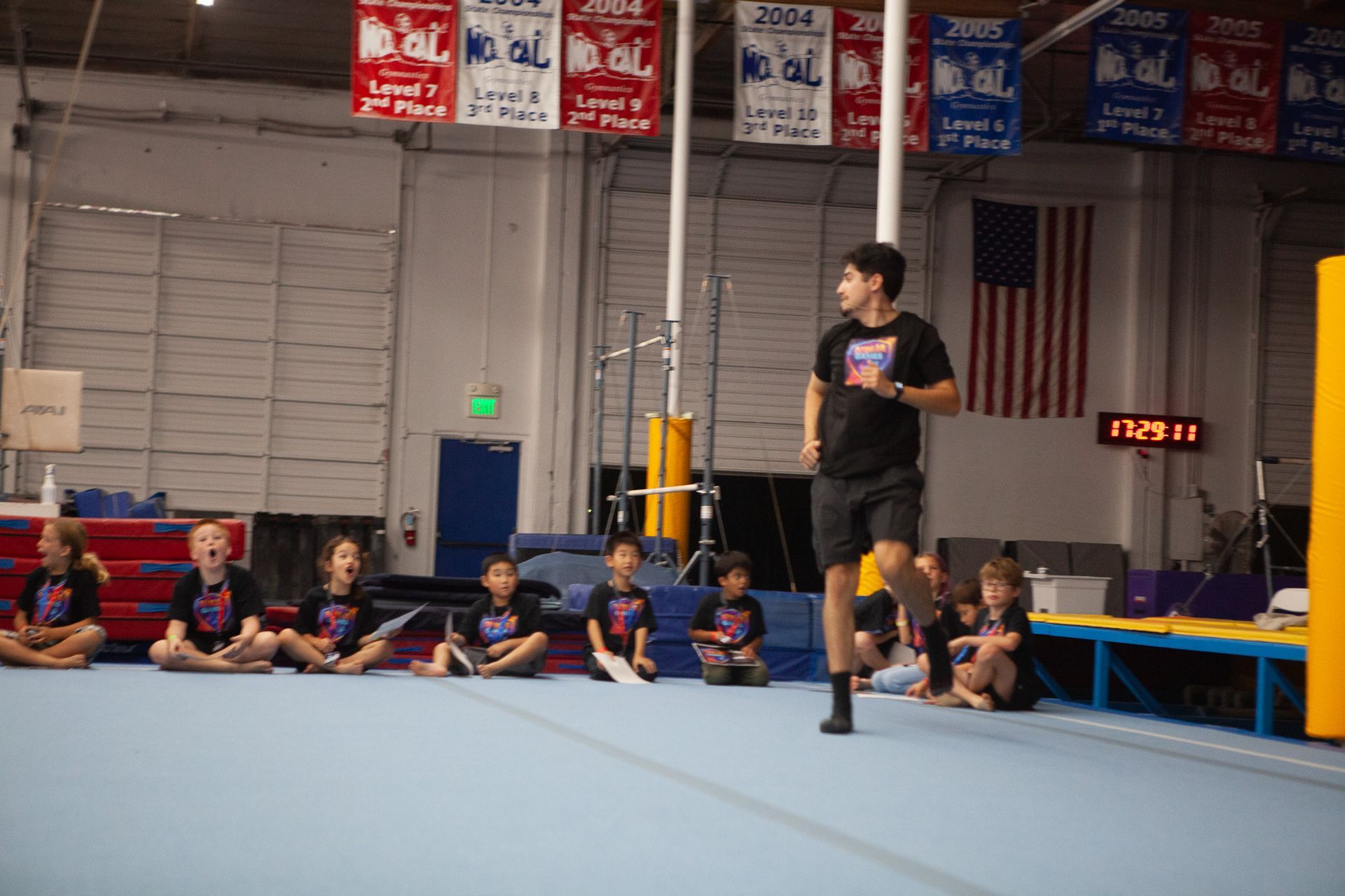 A person runs in a gymnastics gym, facing a seated group of children. An American flag hangs in the background.