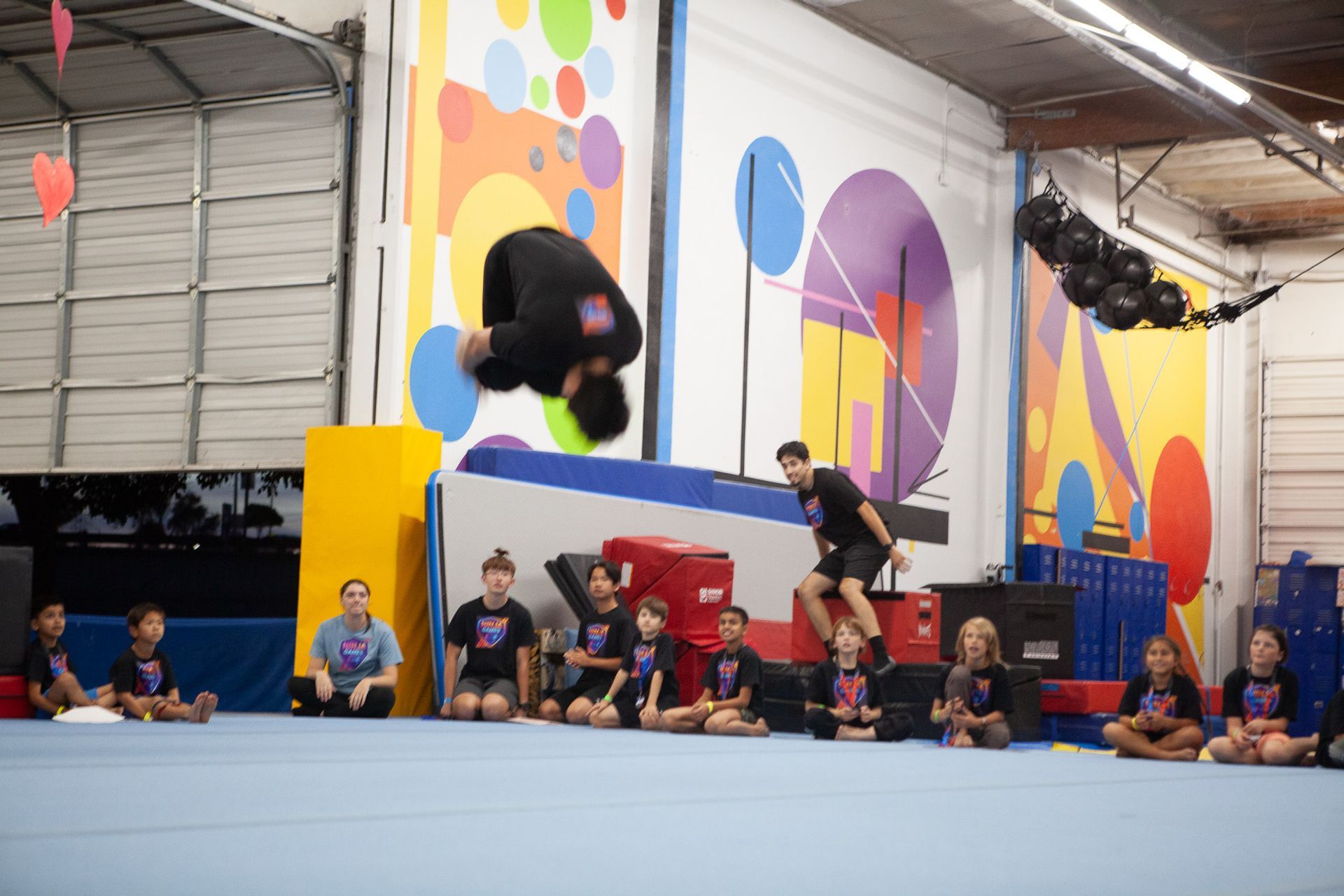Man doing a backflip at a gym. Children watch. Colorful mural in the background.