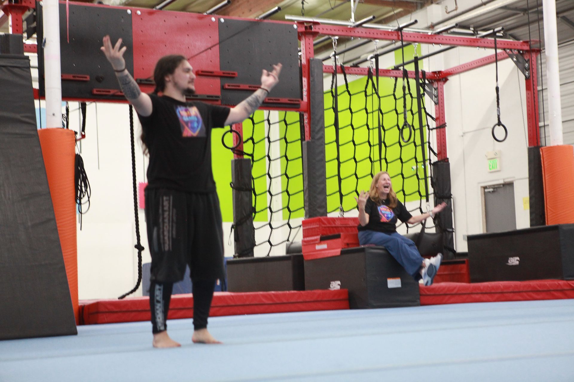 Man with tattoos, arms raised, barefoot, facing a woman seated on a black box, both in a gym setting.