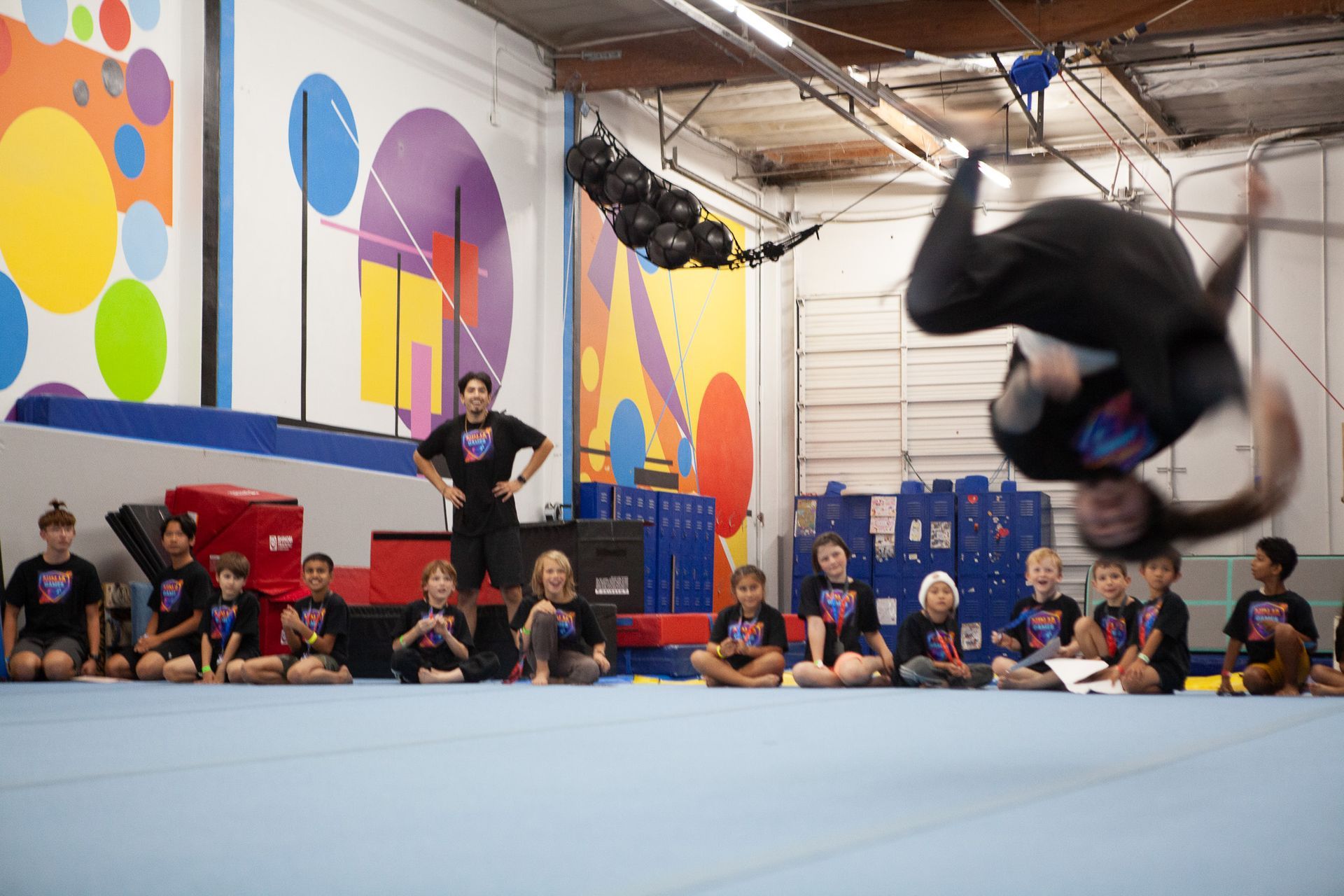 A person doing a backflip in a gym, with kids and an instructor watching.