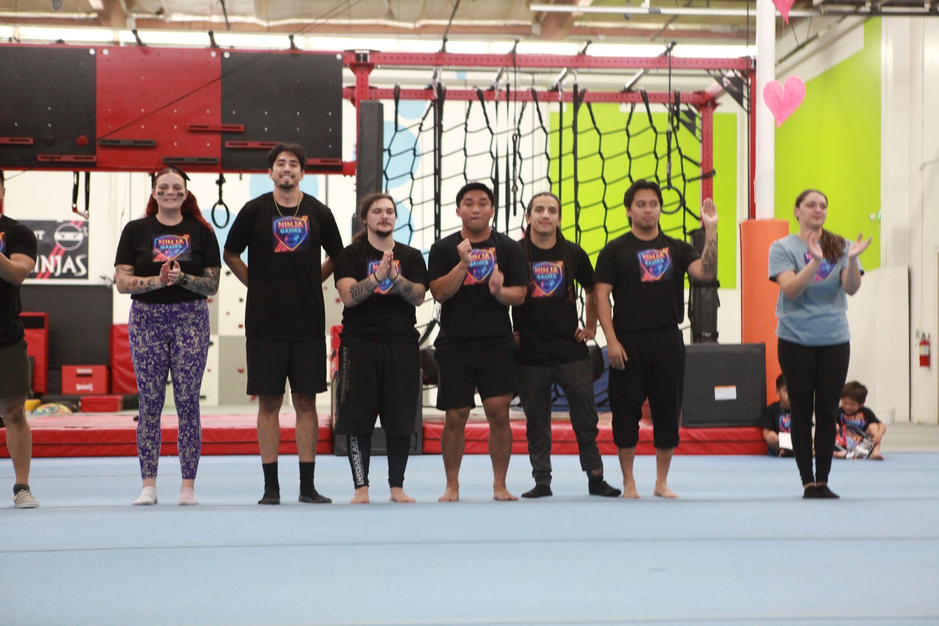 Group of people clapping, standing on a blue mat in a gym. Black shirts, visible obstacle course in background.